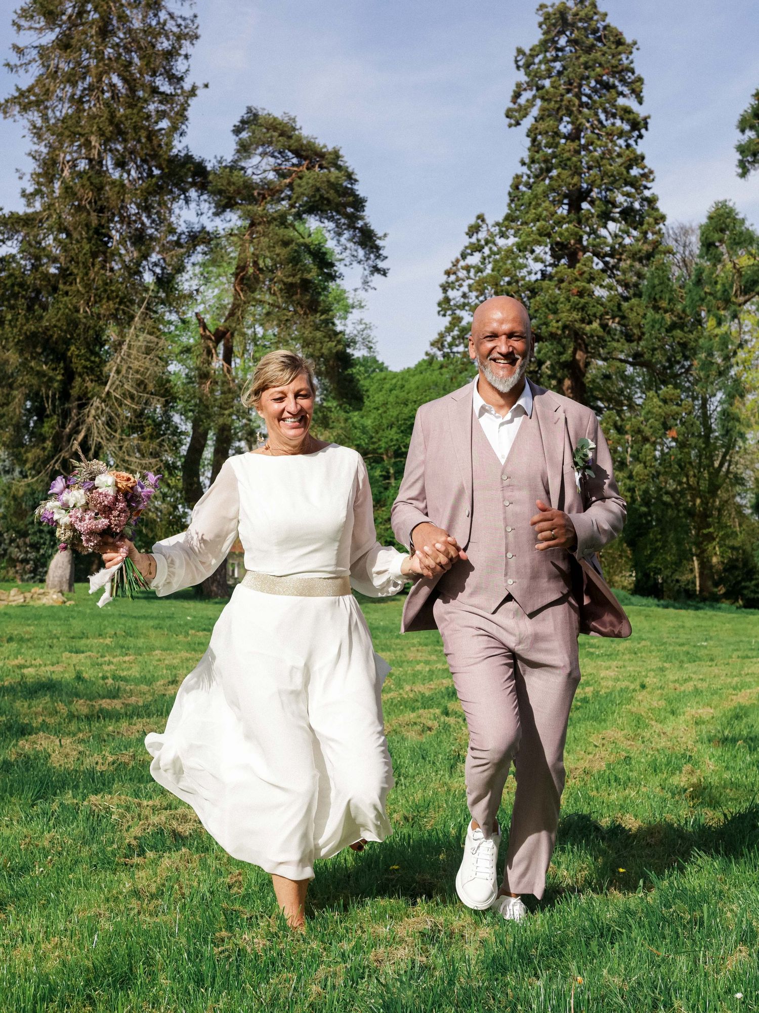 A couple in wedding attire dances and twirls playfully on a grassy hillside with tall trees in the background.