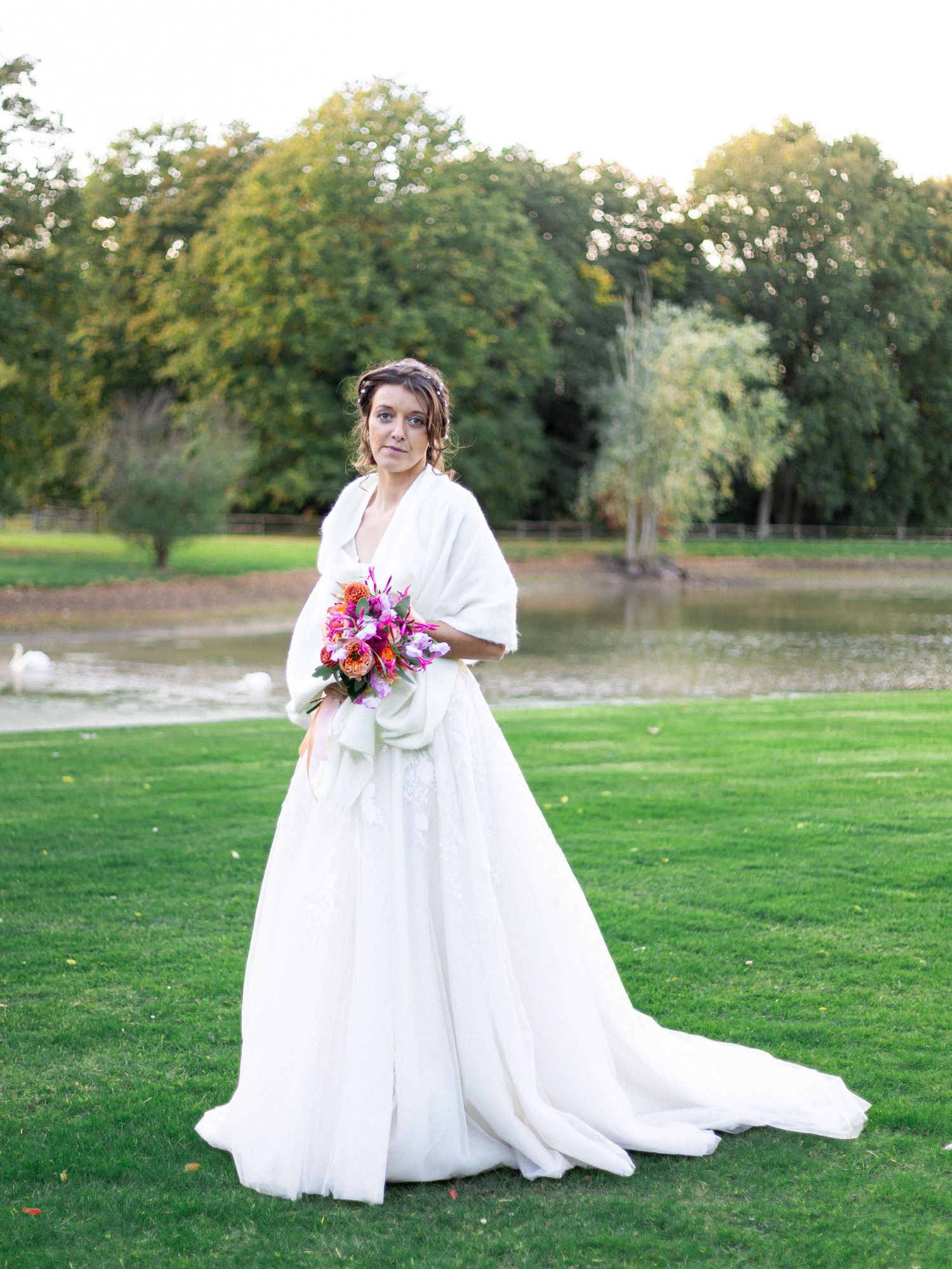 A bride in a flowing white gown holds a colorful bouquet on a green lawn.