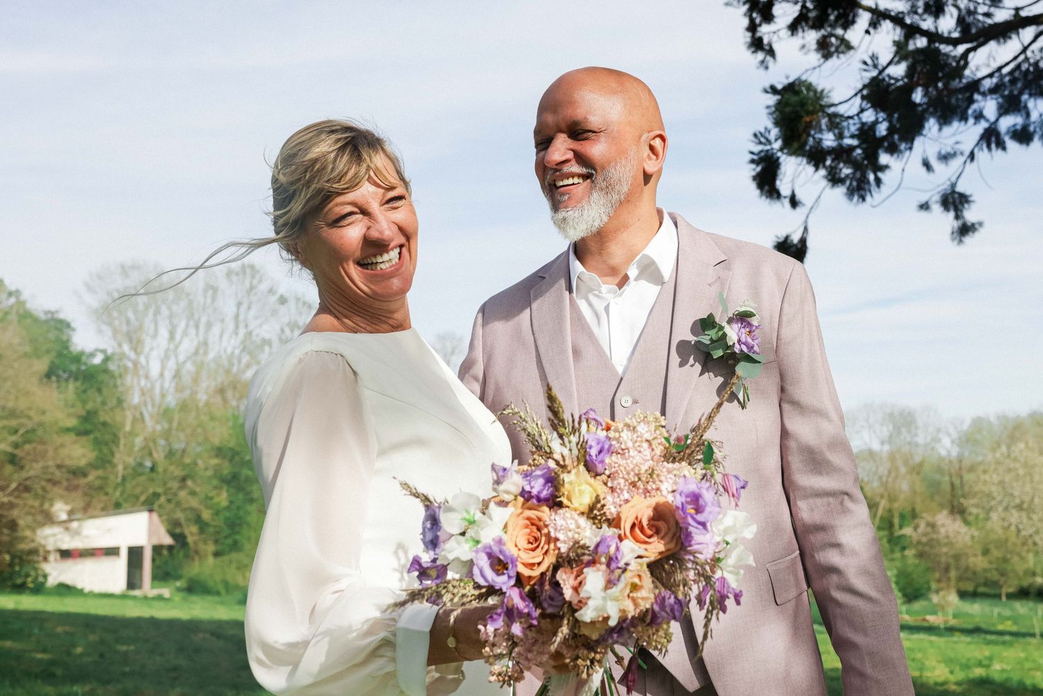 A happy couple in light-colored wedding attire smiles outdoors while holding a vibrant bouquet of pink and purple flowers.