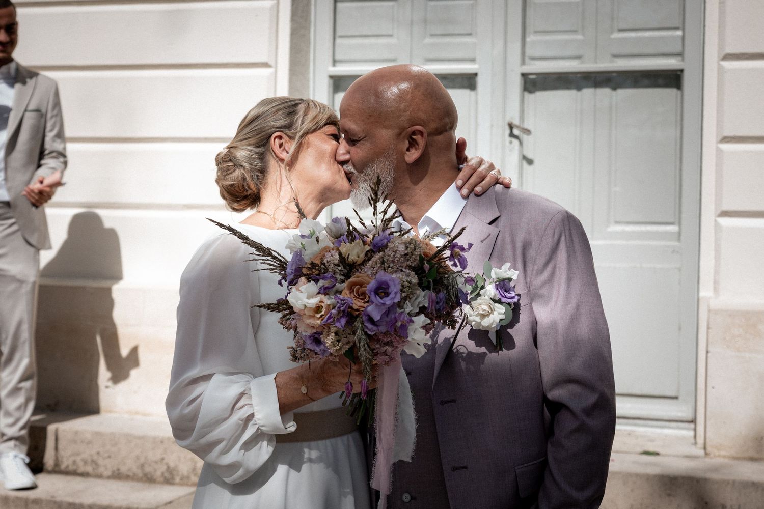 Newlywed couple sharing a romantic kiss outside, bride holding purple and white bouquet, groom in gray suit.