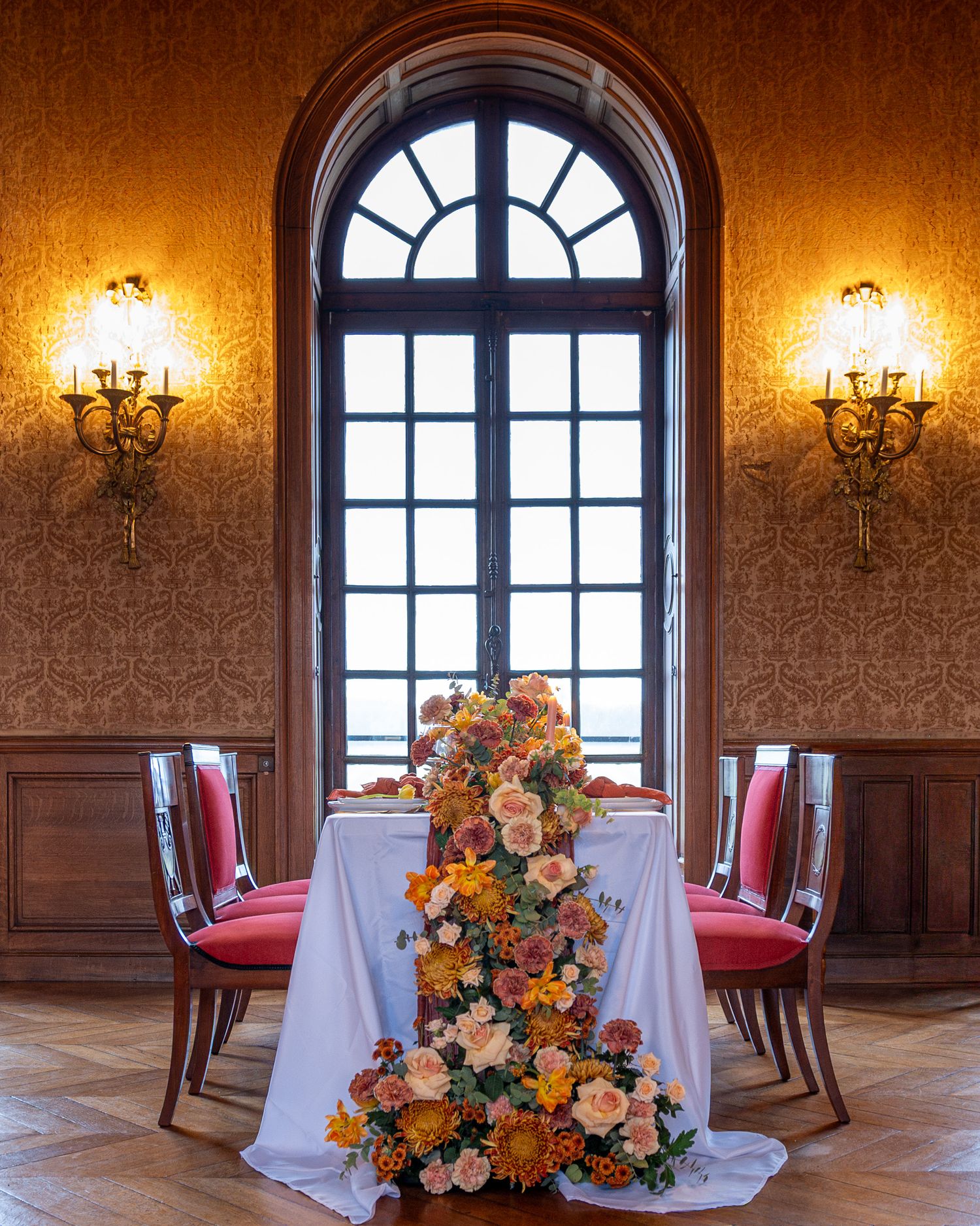 Elegant dining room interior with arched windows, wall sconces, and round tables set with white linens and floral centerpieces.