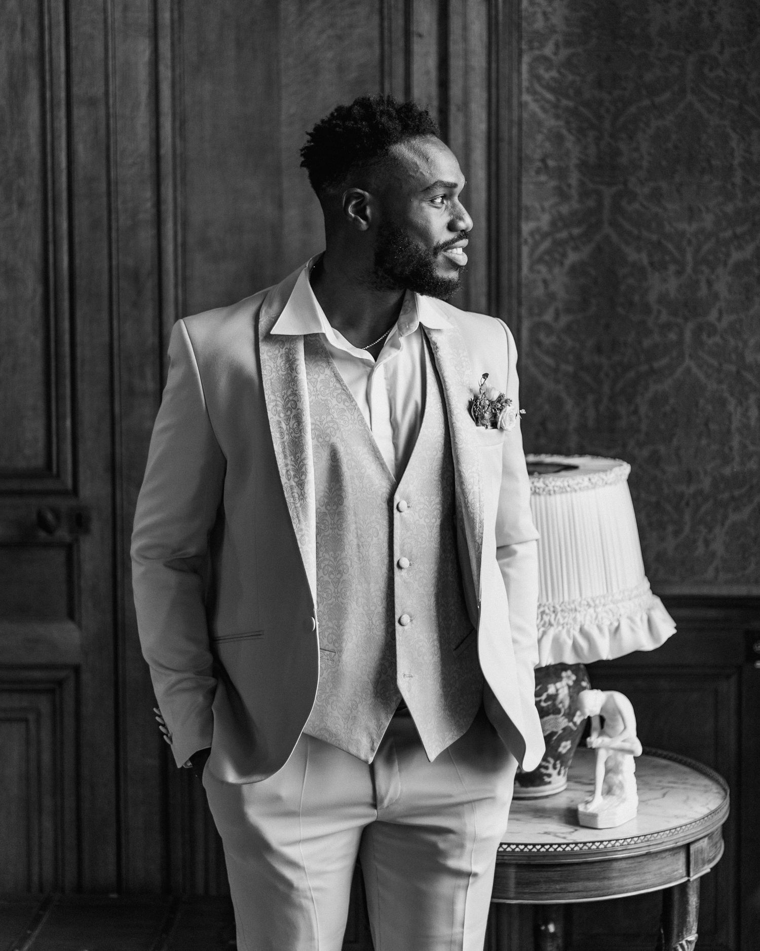 Black and white portrait of groom in white suit standing next to decorative table.