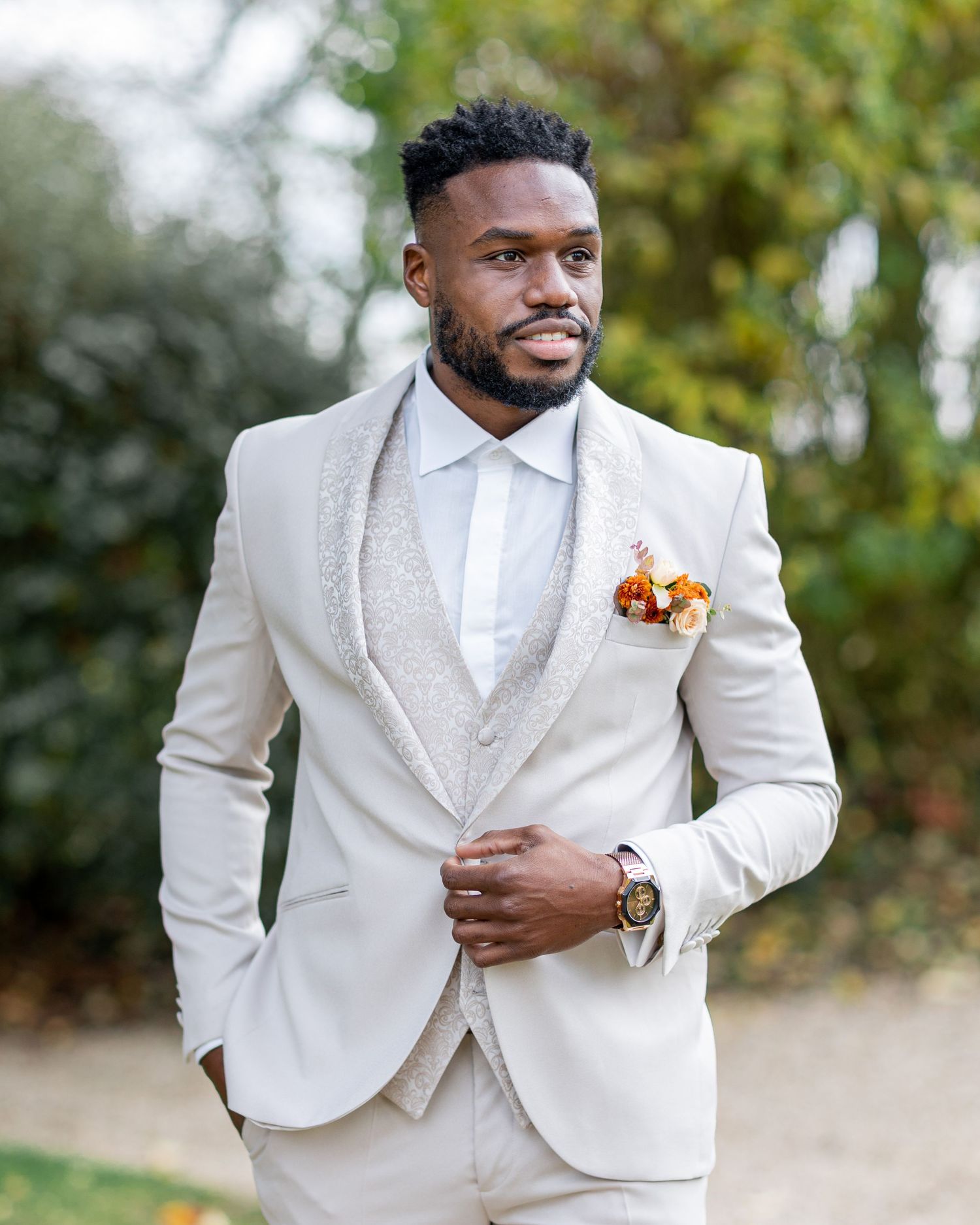 Groom in crisp white tuxedo with floral boutonniere poses outdoors against natural greenery.