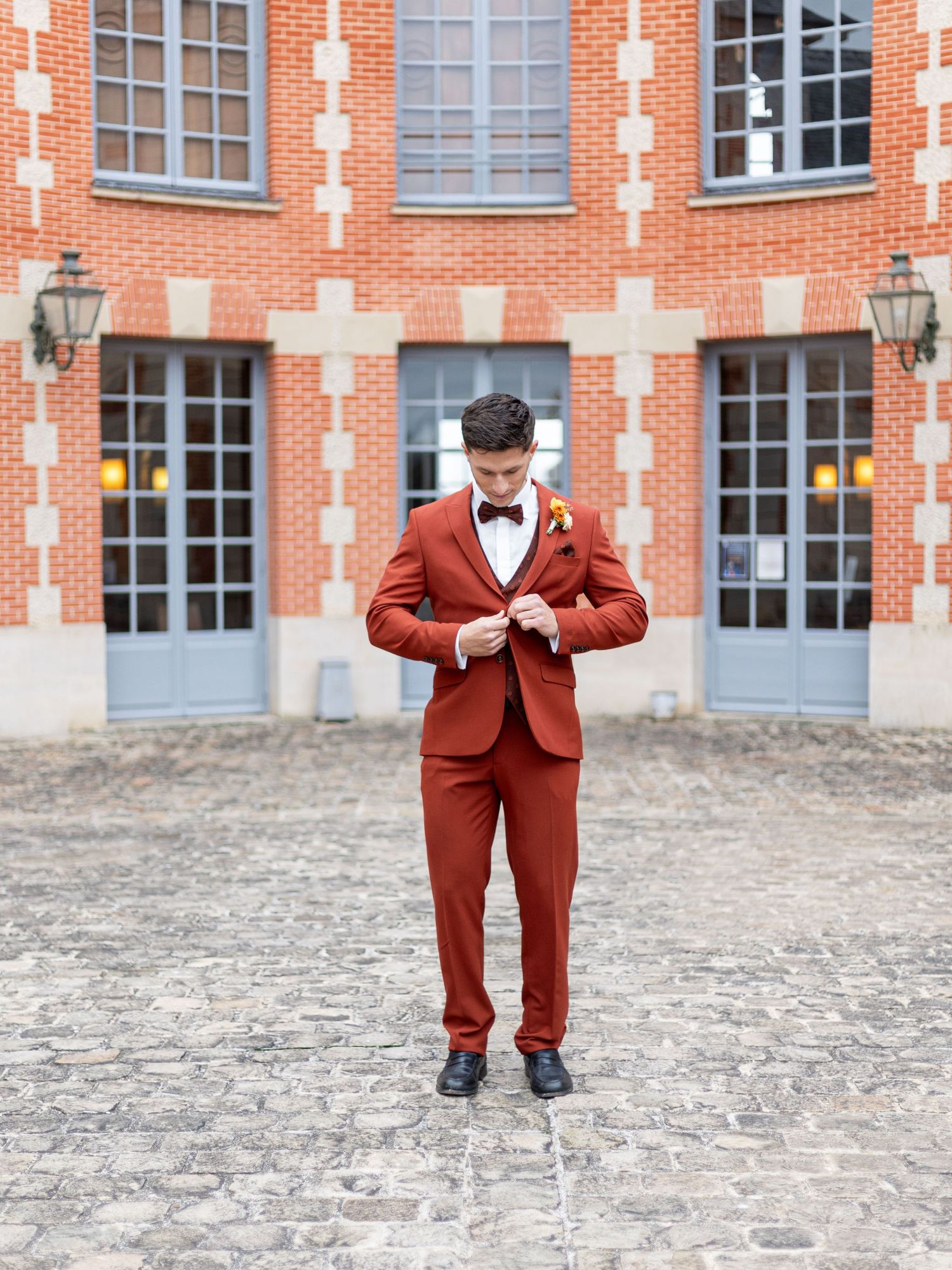 Person in burgundy suit performs playful poses on cobblestone courtyard against brick building.