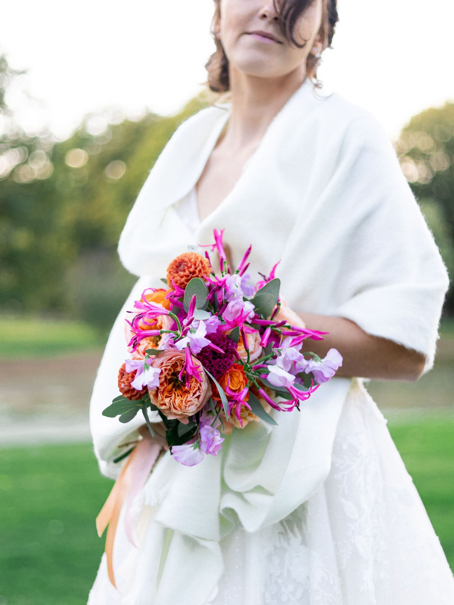 Colorful wedding bouquet with orange and pink flowers held against a white wrap dress outdoors.