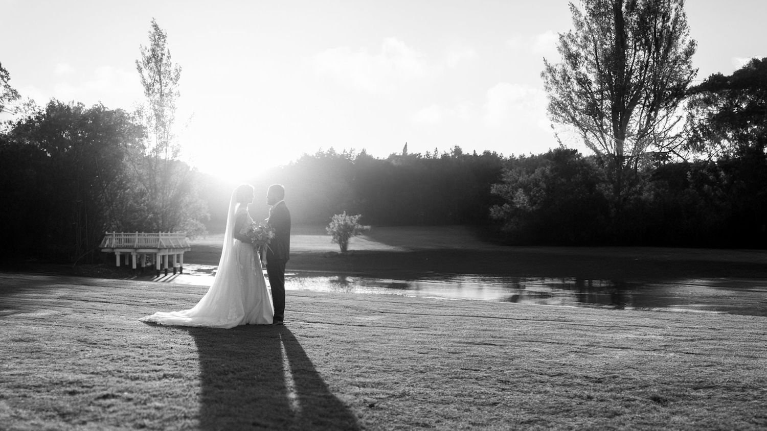Black and white photo of a bride and groom standing by a pond at sunset in a lush garden setting.