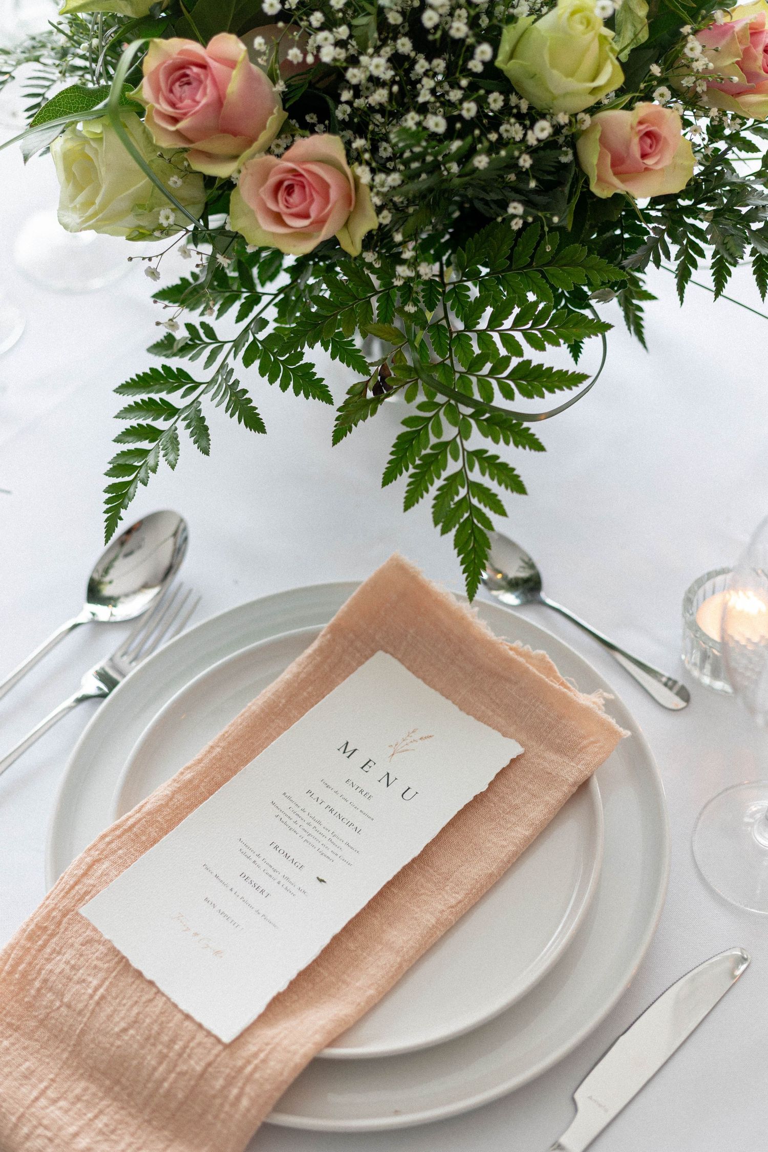 Wedding table setting with peach napkin, white menu card, and fresh floral arrangement.