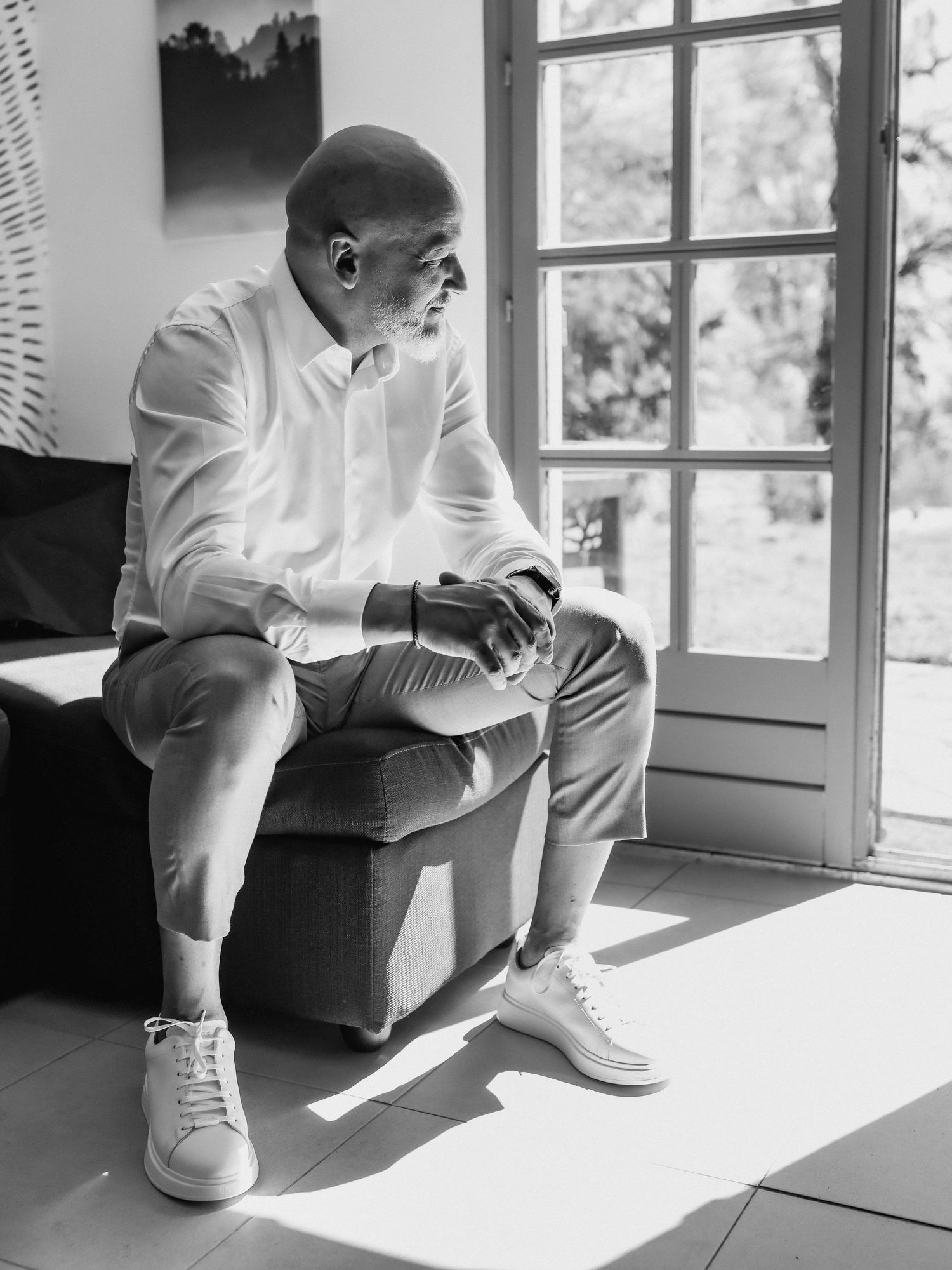 Black and white photo of someone in white clothing sitting pensively by a window, with natural light streaming in.