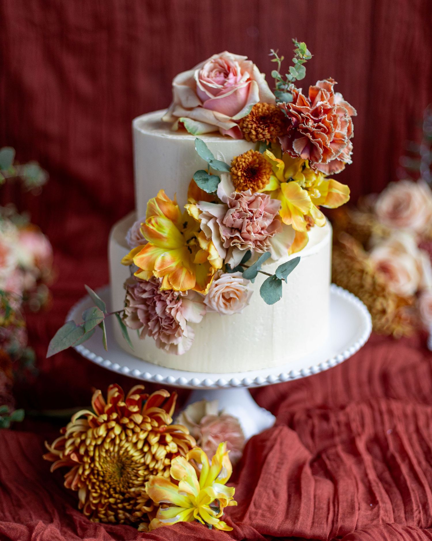 White wedding cake decorated with pink, yellow and orange sugar flowers on burgundy velvet tablecloth.