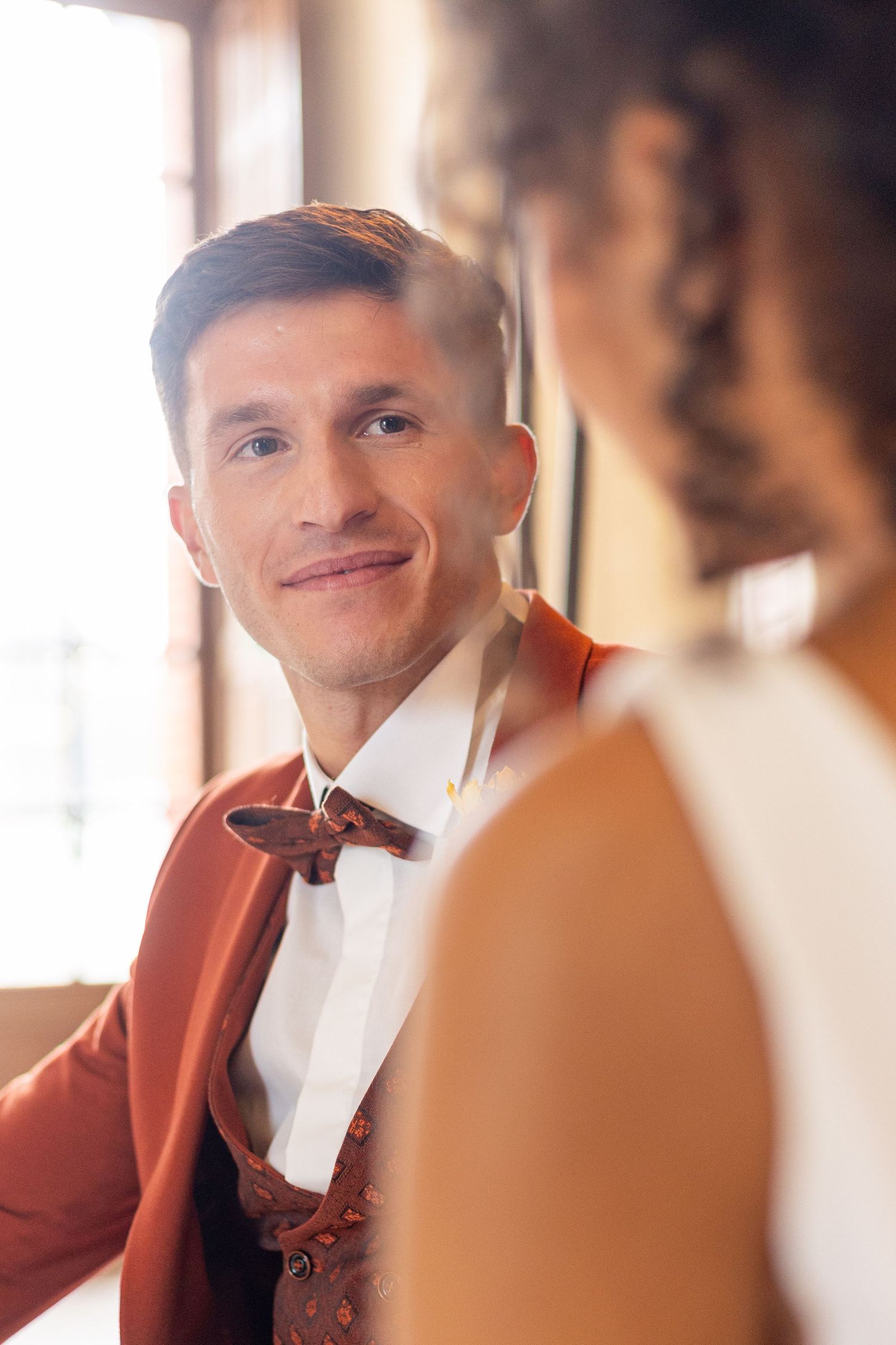 Groom wearing a burgundy tuxedo jacket and bowtie smiles warmly in an elegant indoor setting.