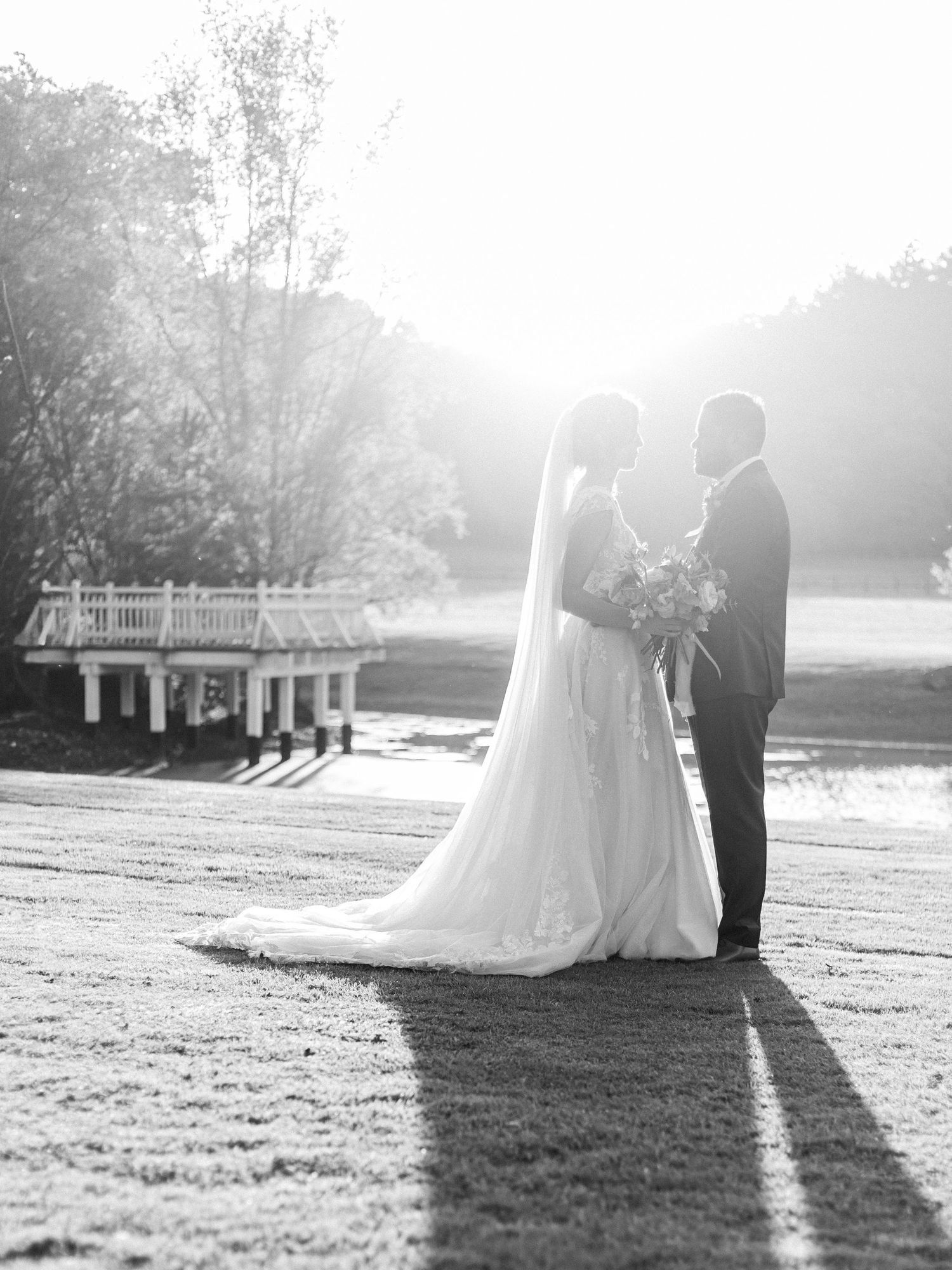 A wedding couple silhouetted against sunlight near a lakeside dock.