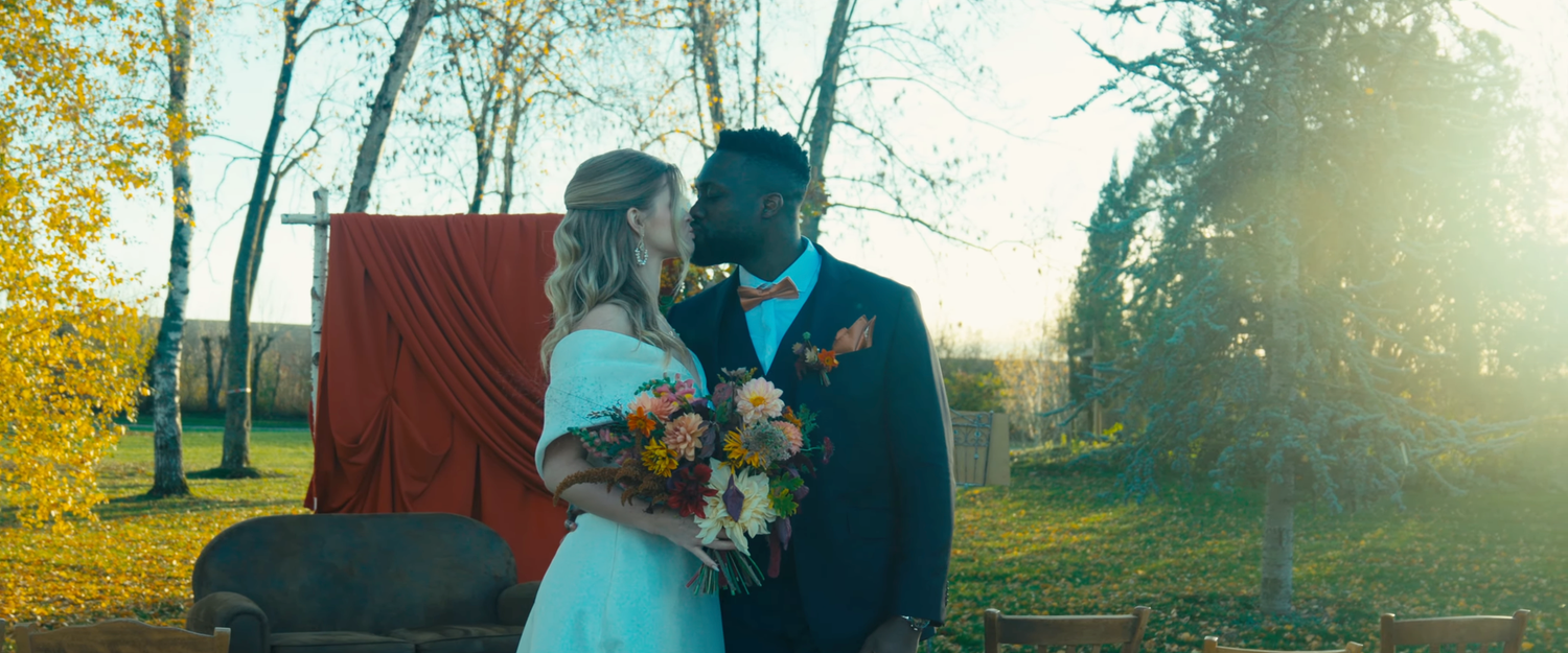 A couple shares a romantic moment in a sunlit autumn setting with golden trees and a red backdrop.