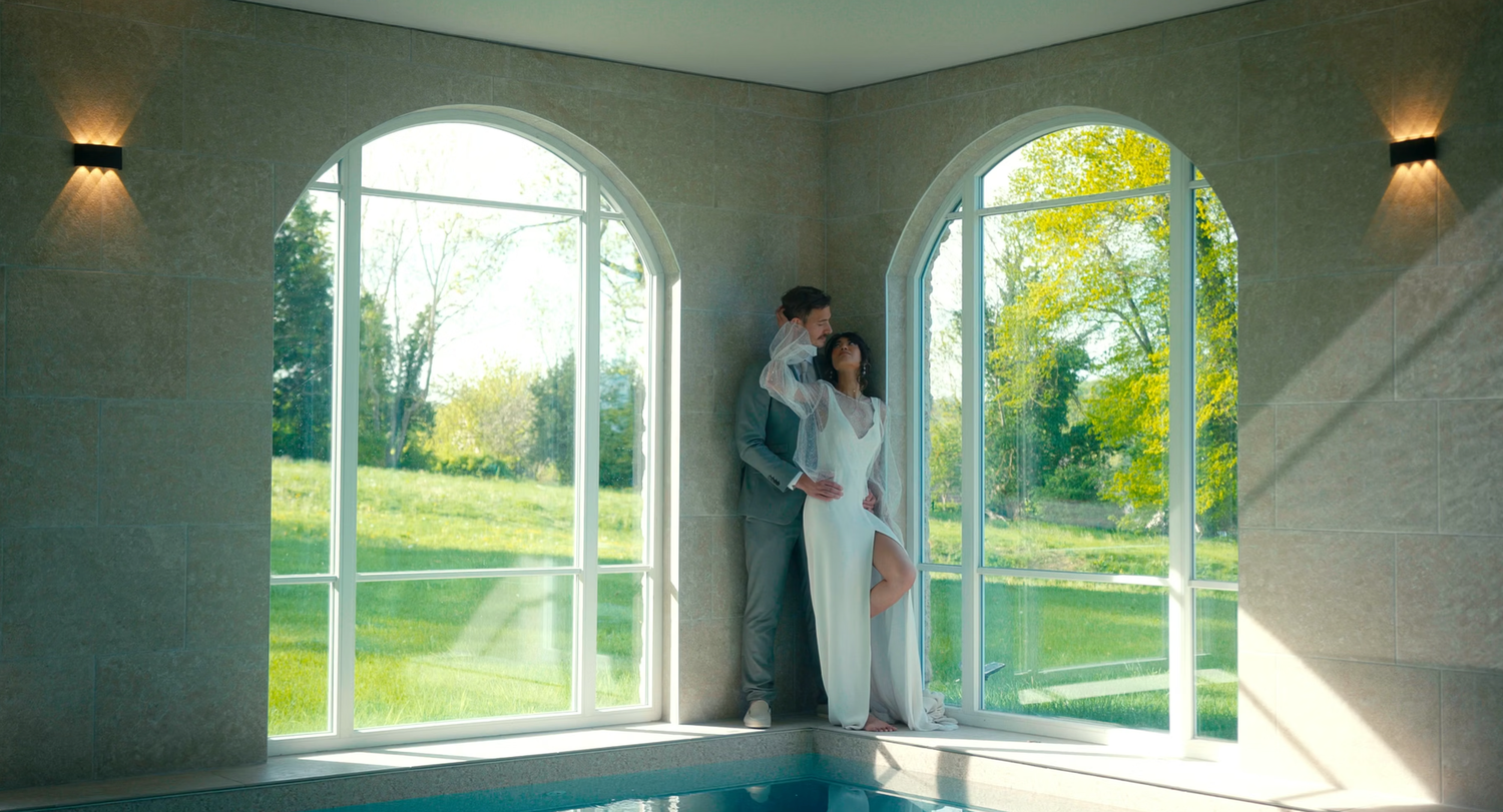 Couple standing by arched windows overlooking a green garden beside an indoor pool in an elegant spa.