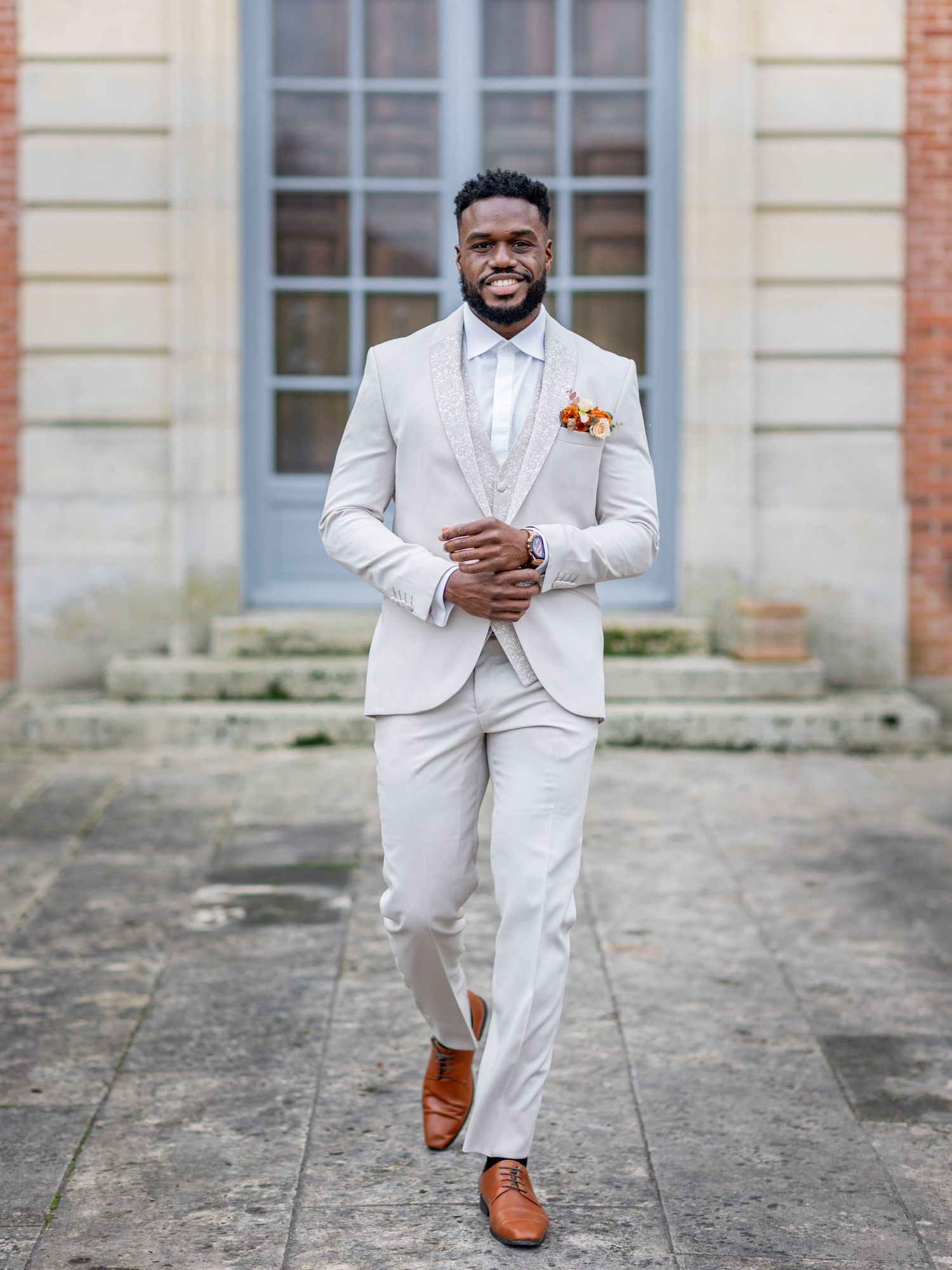 Groom in all-white suit and brown shoes walks confidently across stone courtyard.