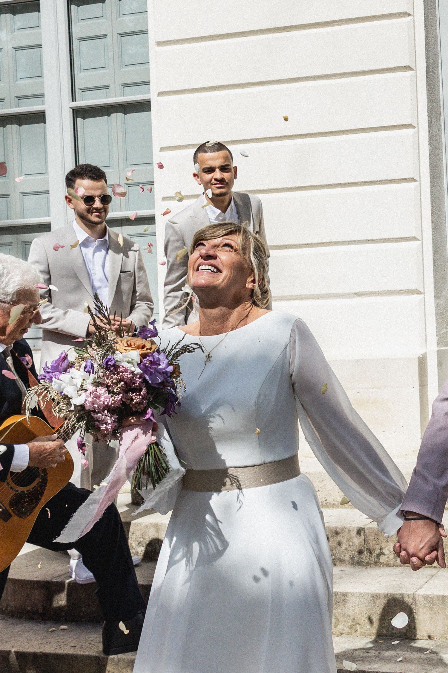 Joyful wedding couple laughing together on outdoor steps with purple floral bouquet and guests in background.