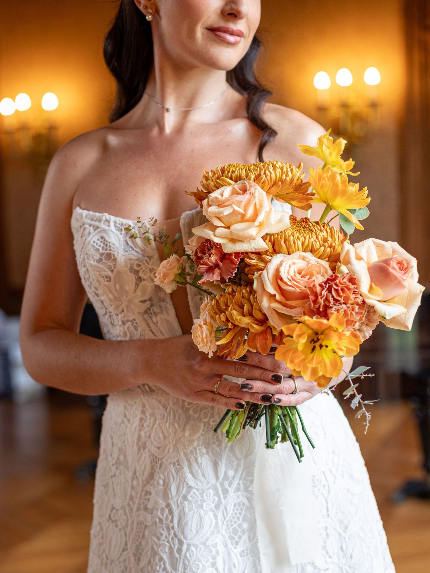 Elegant peach and orange floral wedding bouquet held against a white lace dress in warm lighting.