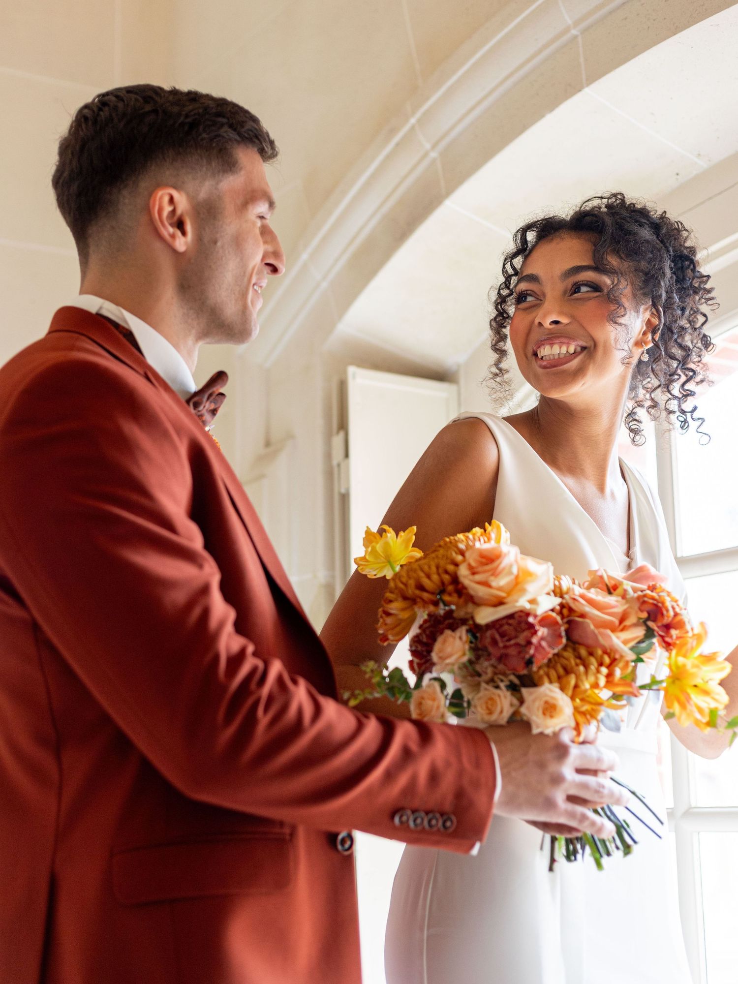 A couple in wedding attire share a joyful moment while holding a vibrant autumn-colored bouquet under an arched doorway.