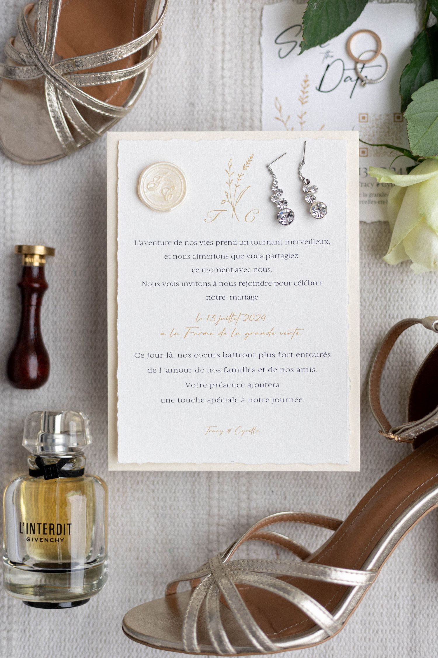 Wedding details flatlay with invitation, perfume bottle, and silver shoes on textured fabric.