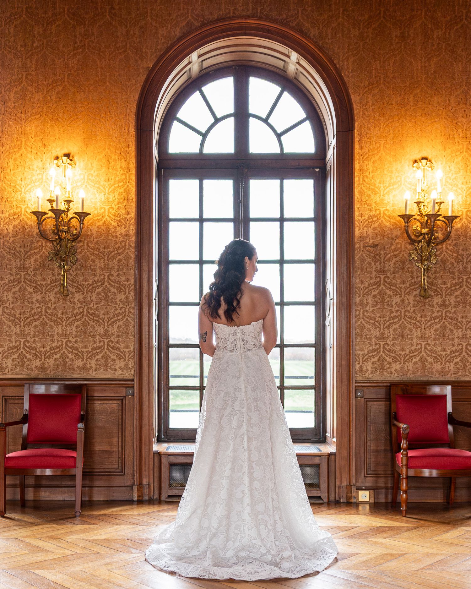 Wedding dress displayed in elegant ballroom with ornate wallpaper and arched windows.