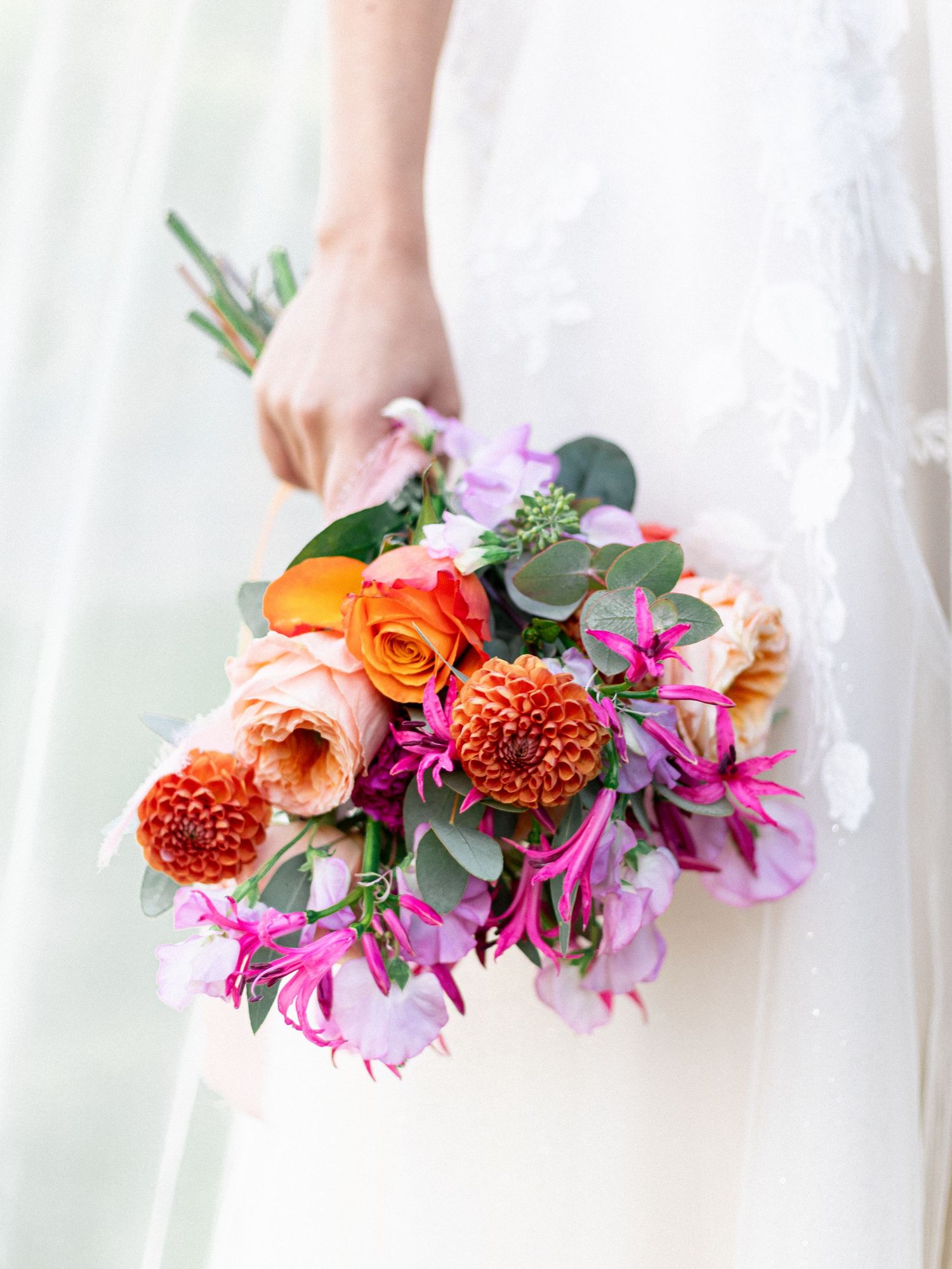 Vibrant bridal bouquet with orange roses, pink dahlias and purple flowers against a white dress background.