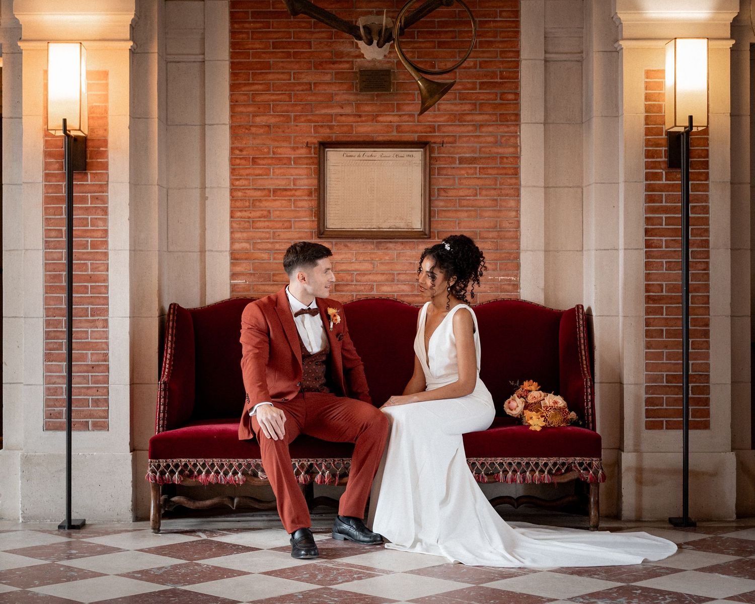 Bride and groom sitting on red velvet sofa in elegant brick venue, groom in burgundy suit, bride in white gown.