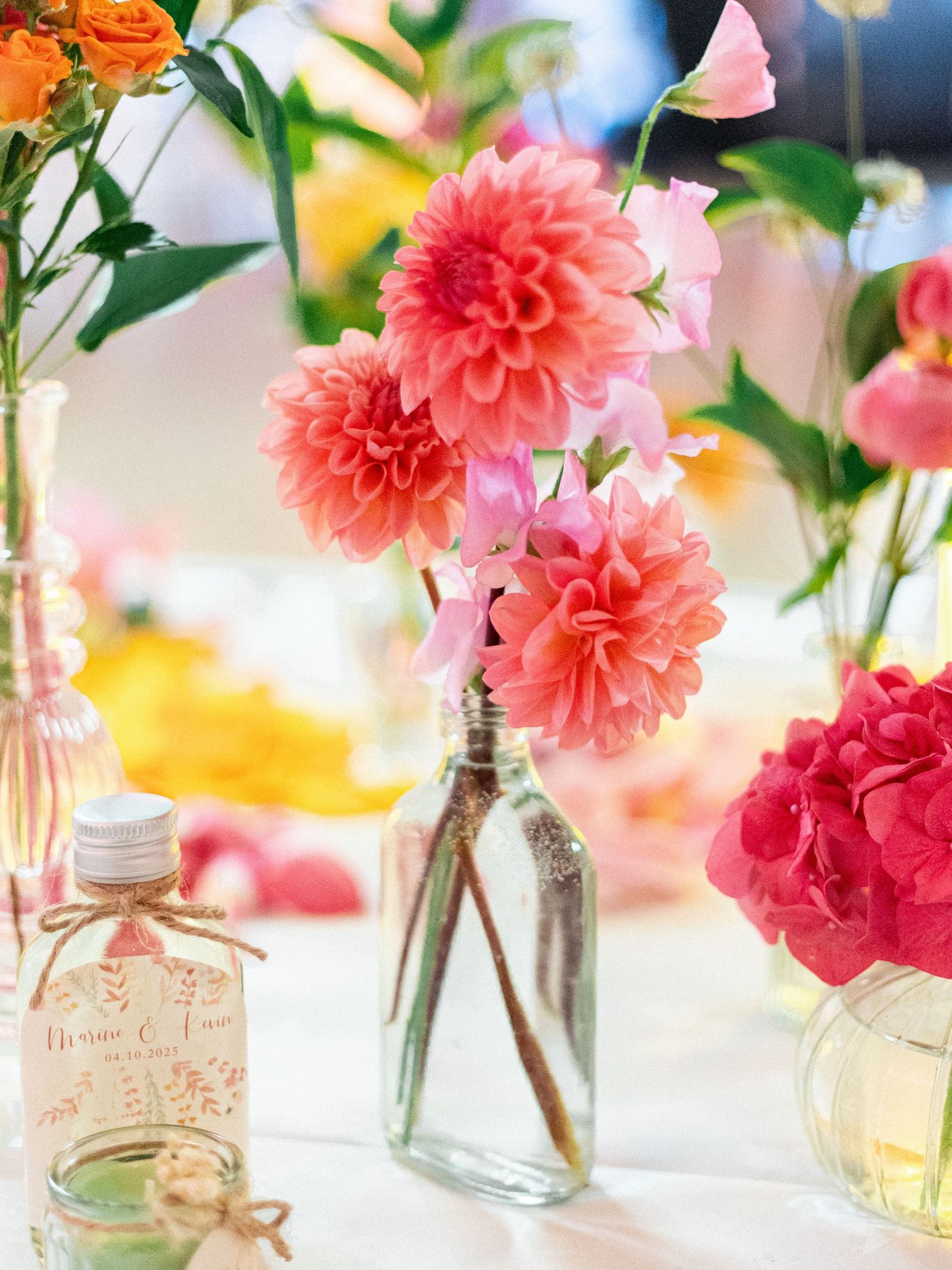 Pink dahlias and coral flowers arranged in clear glass bottles on a white tablecloth.