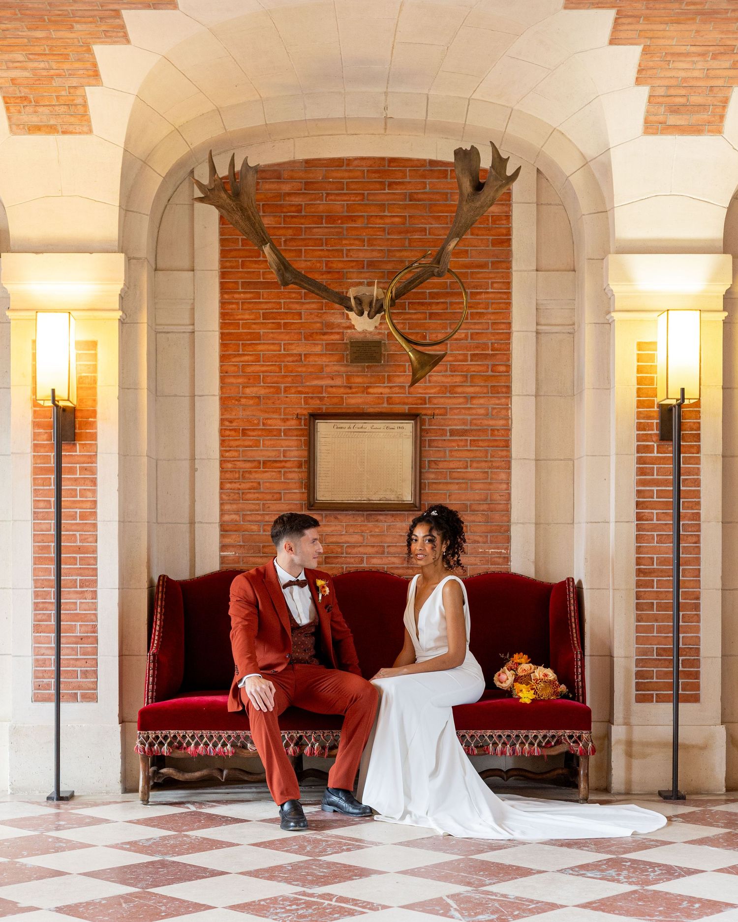 Wedding couple in white dress and burgundy suit posed in grand arched hallway with antler decor.