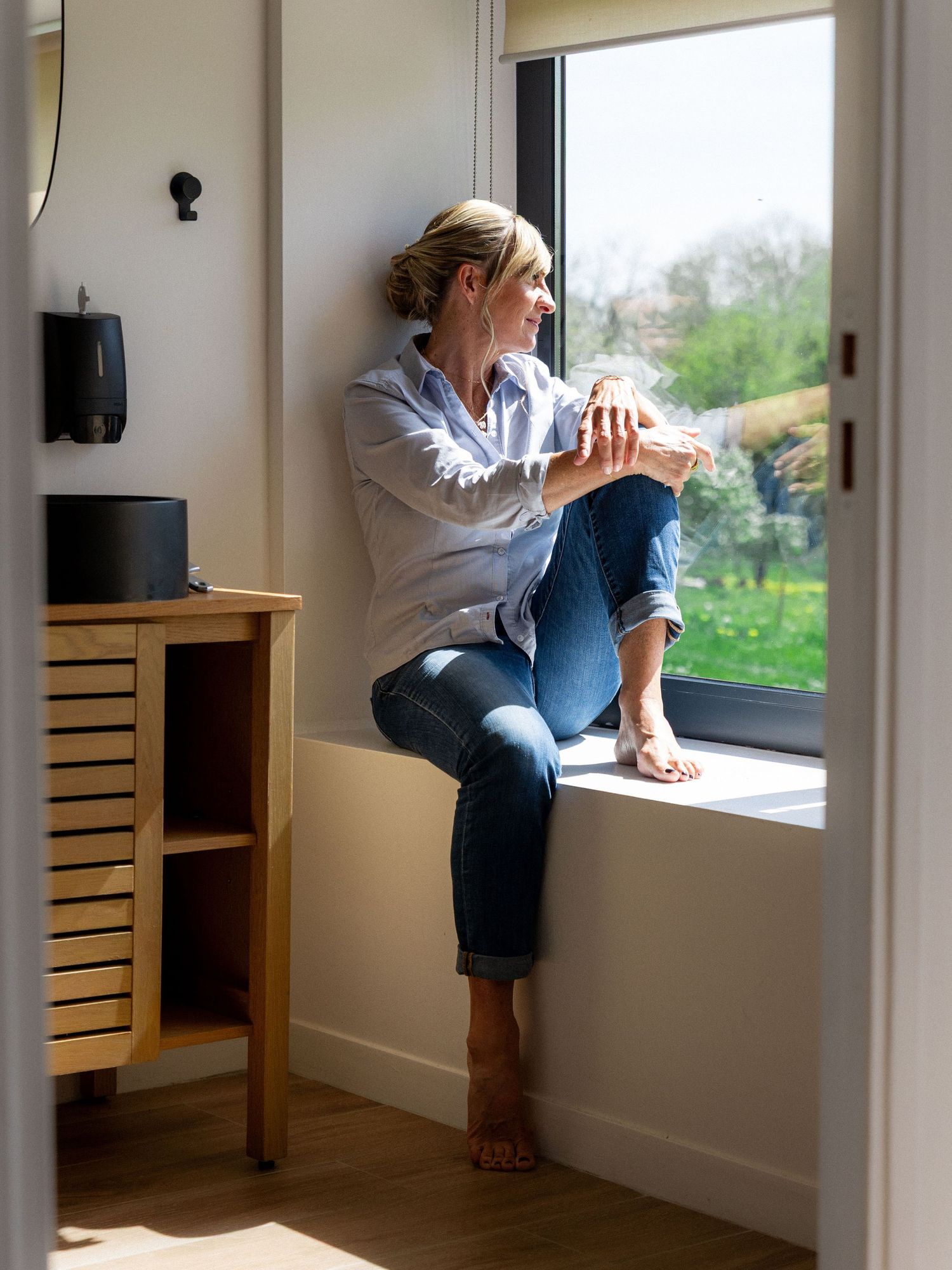 Someone sits contemplatively on a window sill in casual clothing, gazing outside at a garden view on a bright day.