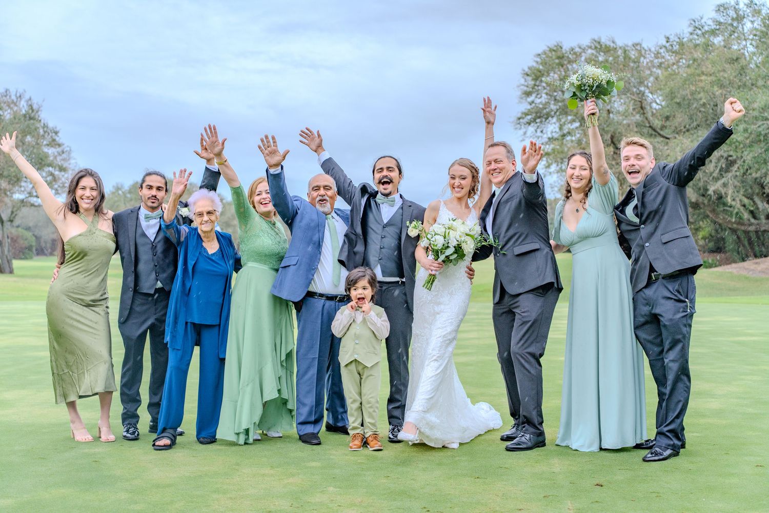 Wedding party celebrates with raised arms in a coordinated photo line on grassy lawn near water.