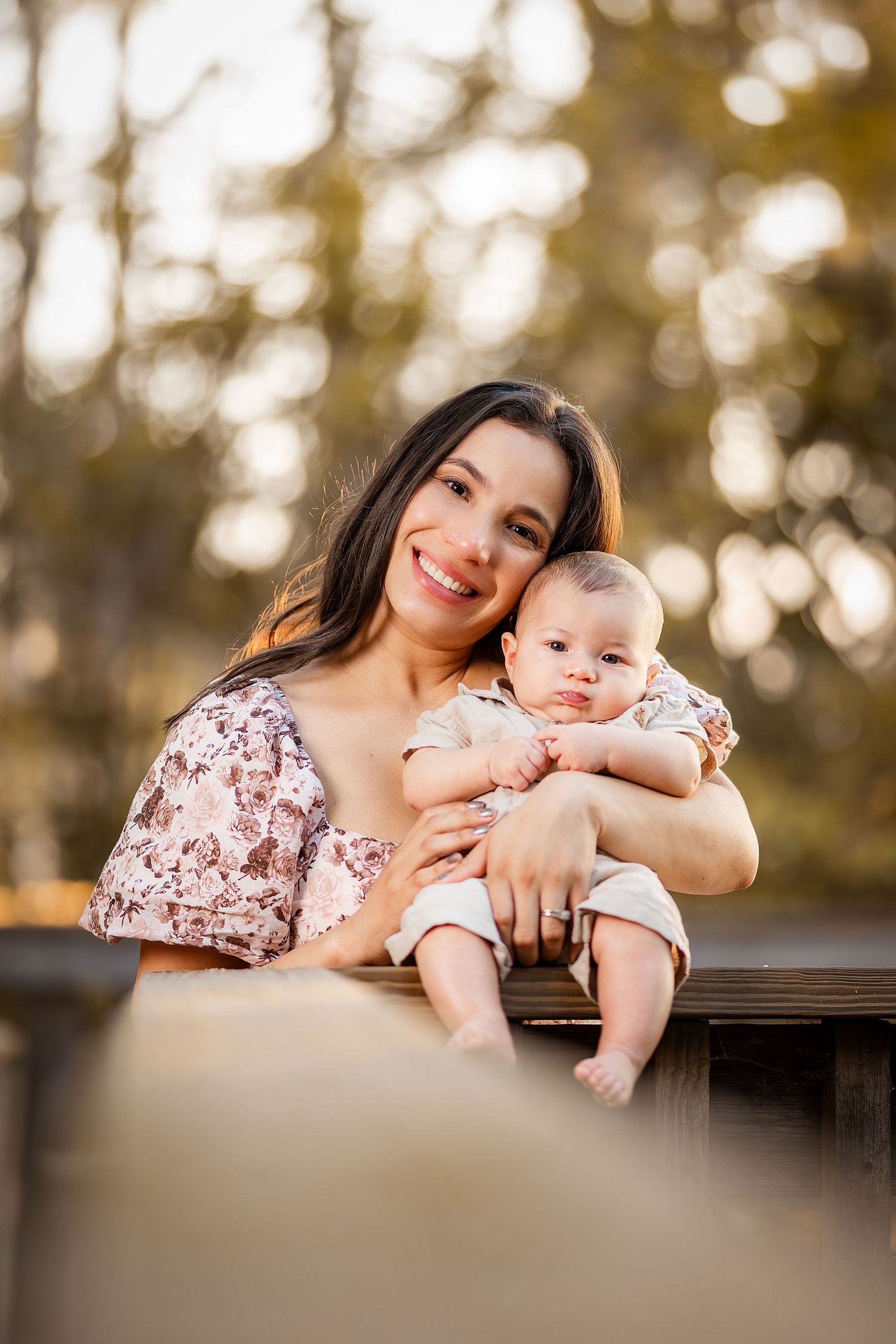 Mother in floral dress holds and cuddles with baby on wooden deck railing during golden sunset.