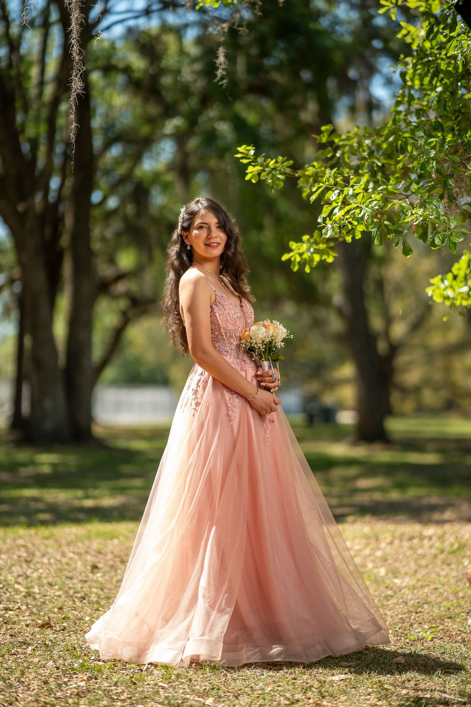 Young woman in pink tulle ball gown holding bouquet, posing outdoors among lush green trees in sunlight.