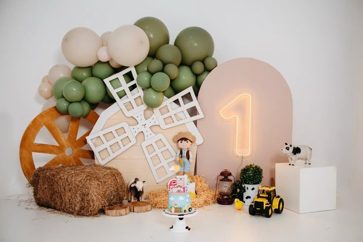Farm themed first birthday setup with windmill, hay bales, and tractor toys against balloon arch backdrop.