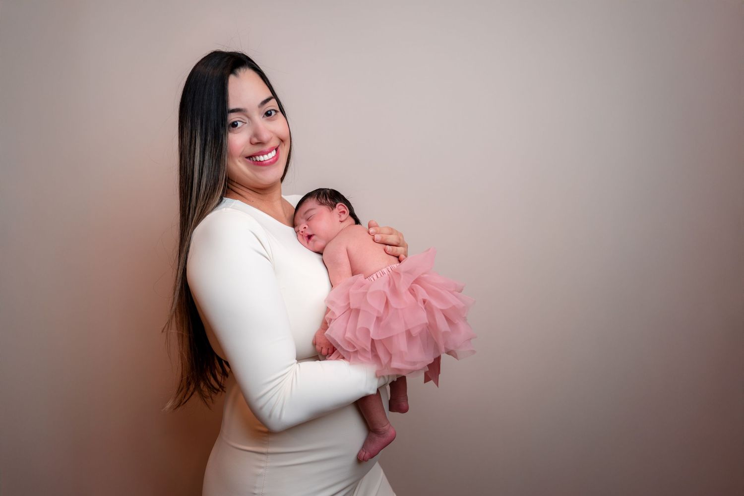 Parent in white dress holds sleeping newborn wearing fluffy pink tulle against neutral backdrop in studio portrait.