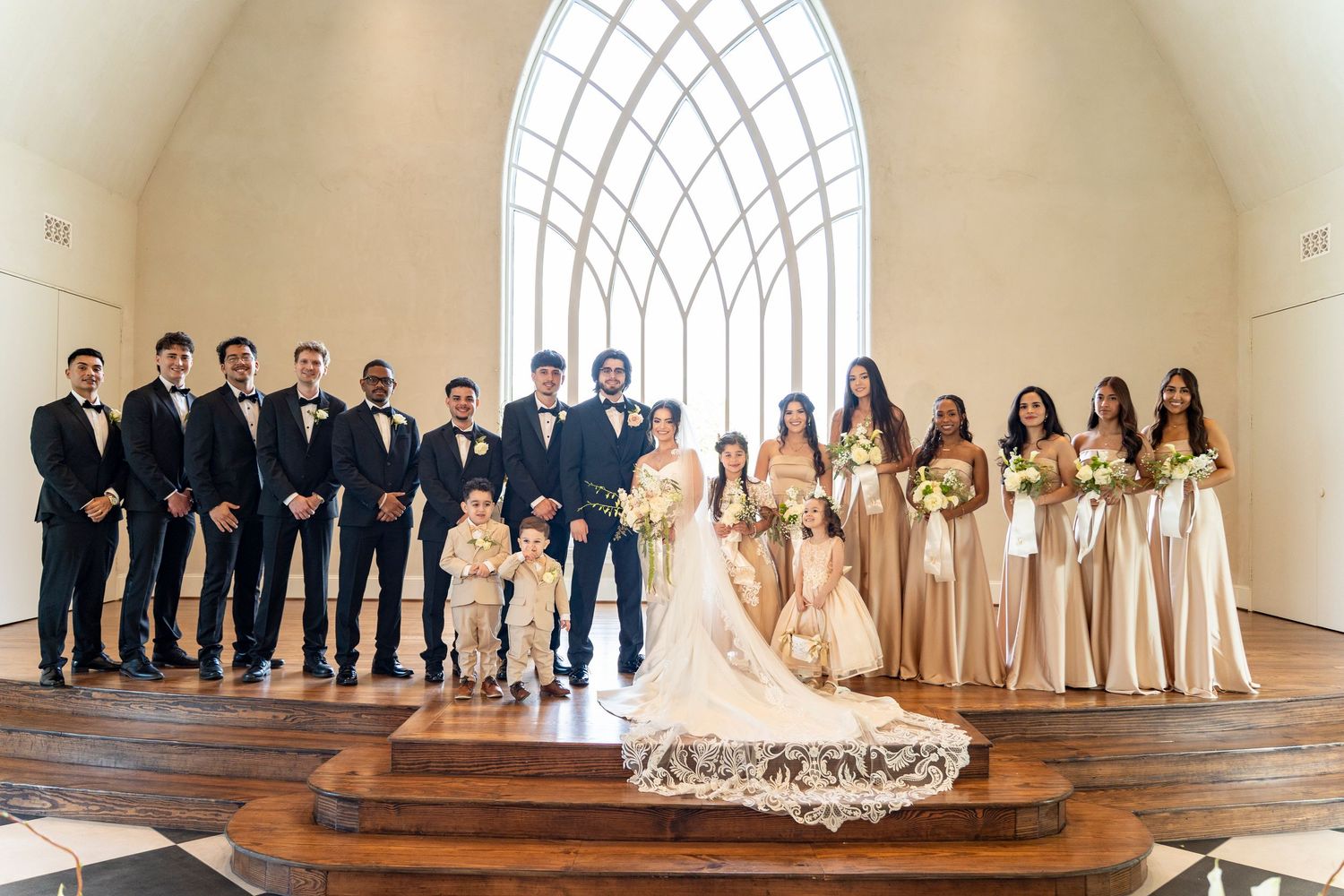 Wedding party poses in white chapel with arched window, bridesmaids in champagne gowns and groomsmen in tuxedos.