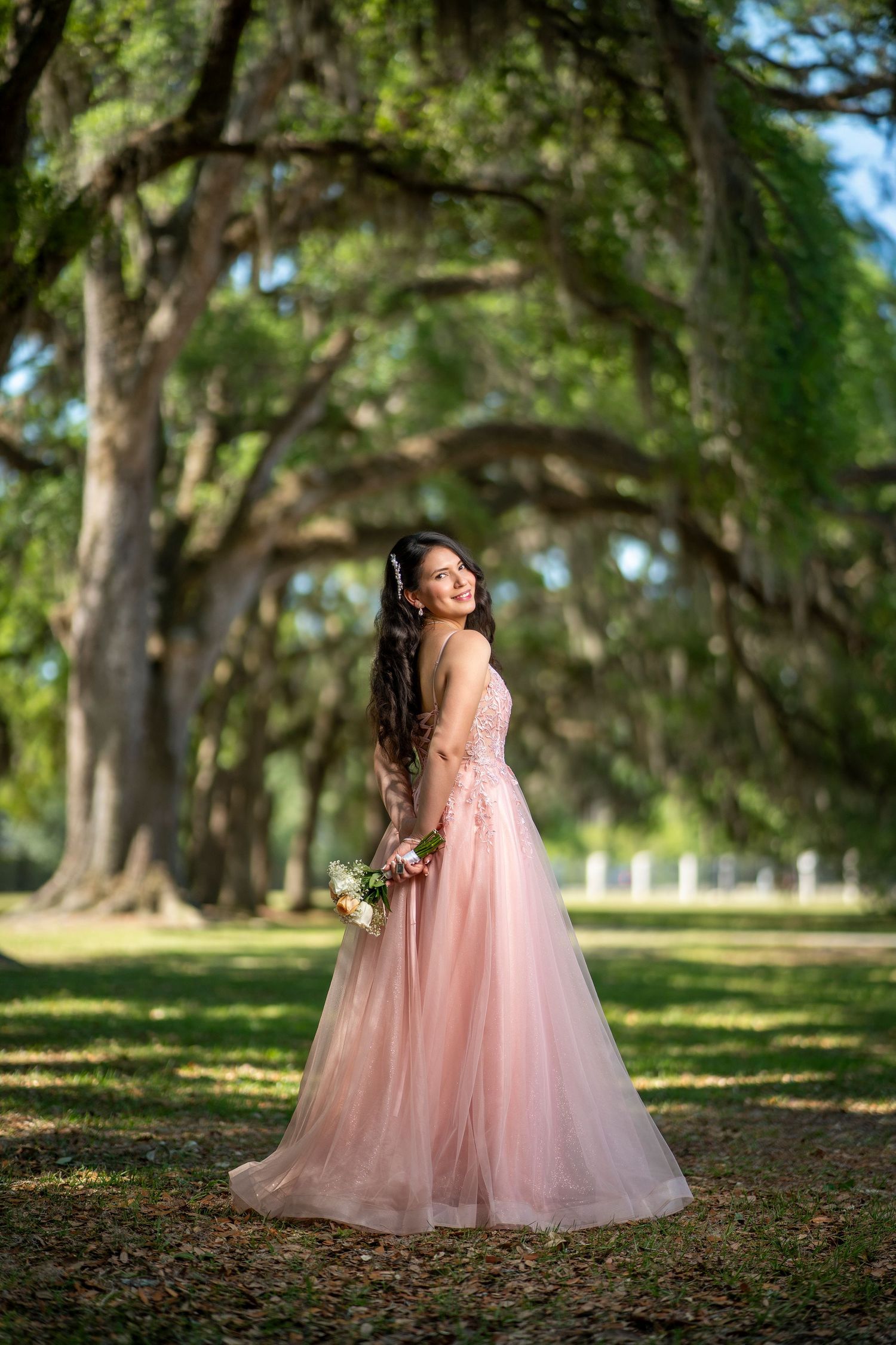 A woman in a pink tulle ball gown holds a bouquet under moss-draped oak trees in a lush green park.