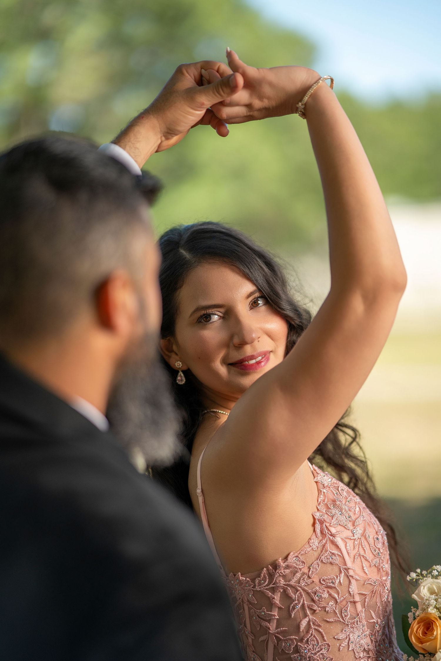 Bride in pink lace gown dancing with groom outdoors, arms raised, smiling elegantly at a wedding celebration.