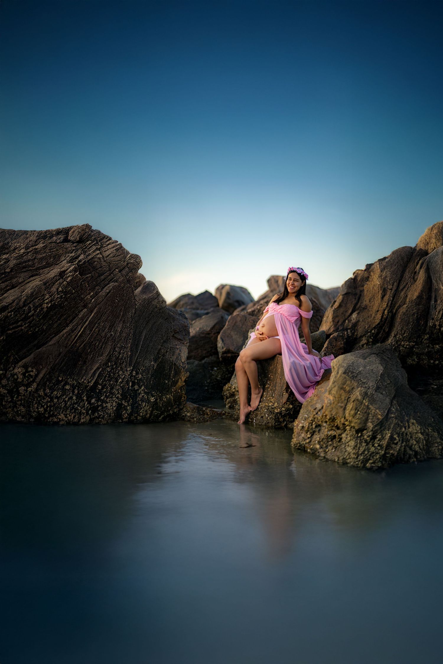 A figure in a flowing pink dress sits gracefully on rocky coastal formations at sunset with calm waters below.