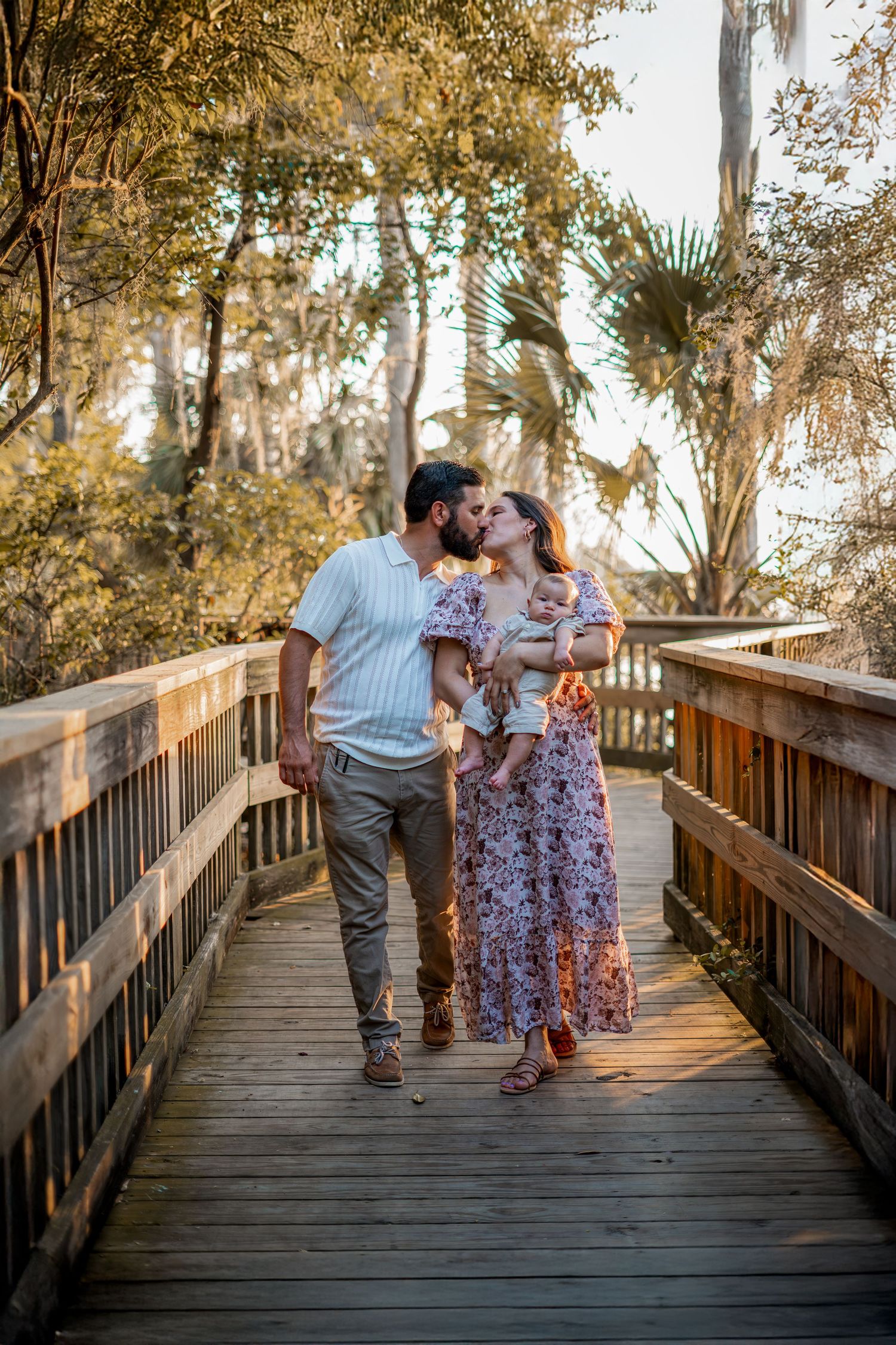 A couple shares a romantic moment while walking together on a wooden boardwalk surrounded by trees at sunset.
