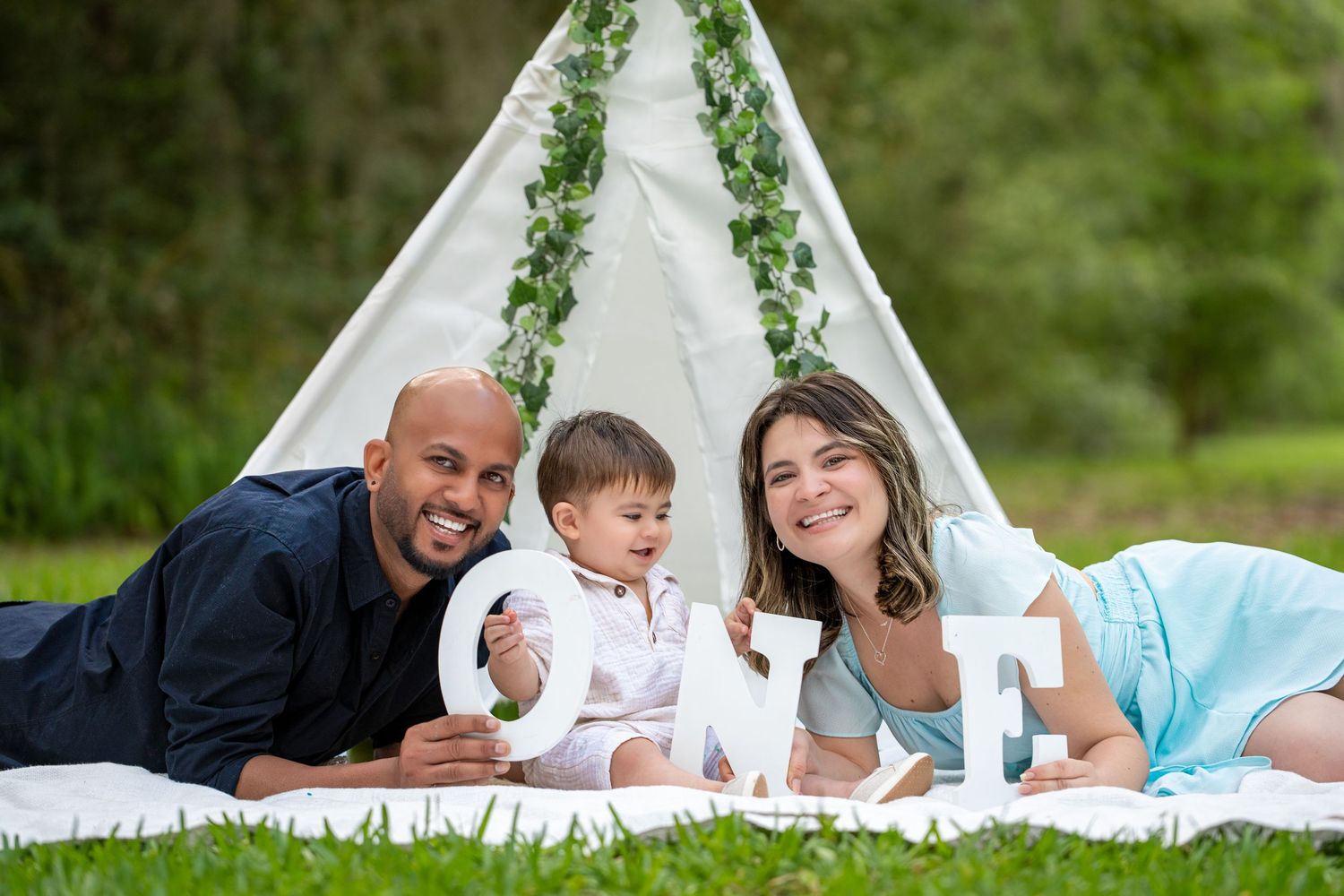 Family lounges together on white blanket with decorative teepee tent and greenery in outdoor setting.