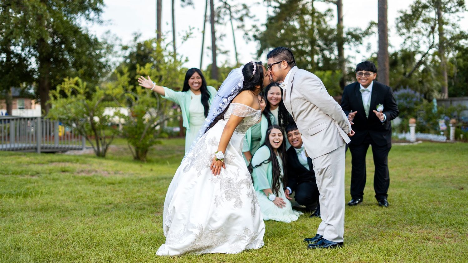 Bride and groom sharing a kiss outdoors while wedding guests celebrate and pose joyfully behind them on green grass.
