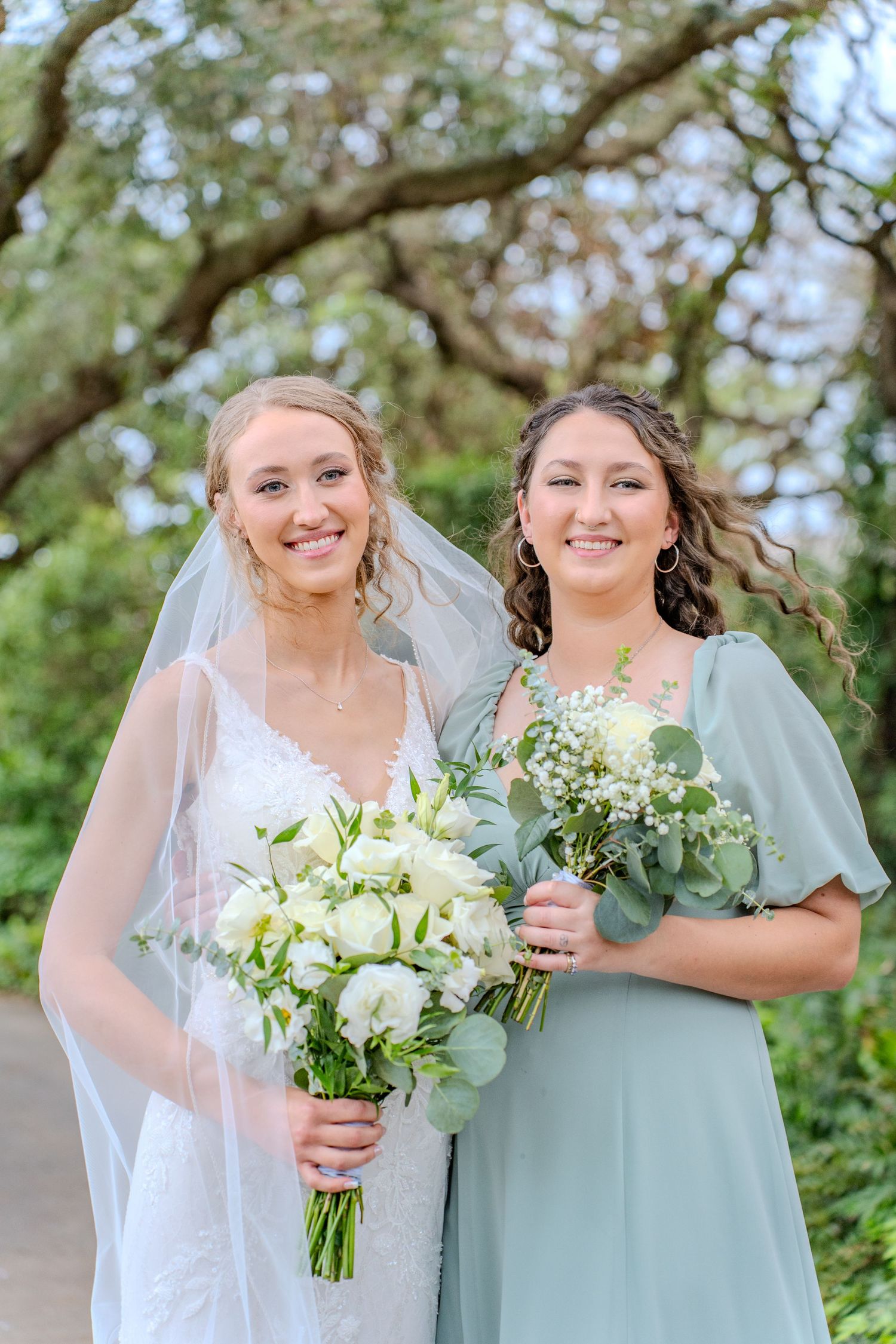 Wedding party members smile together outdoors while holding white floral bouquets in mint green and white attire.