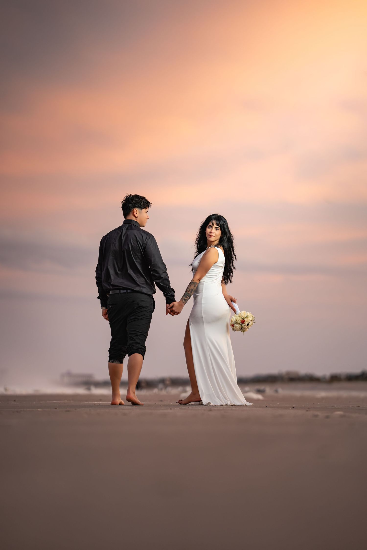 A newlywed couple walks along a sandy beach at sunset with the bride holding a bouquet.