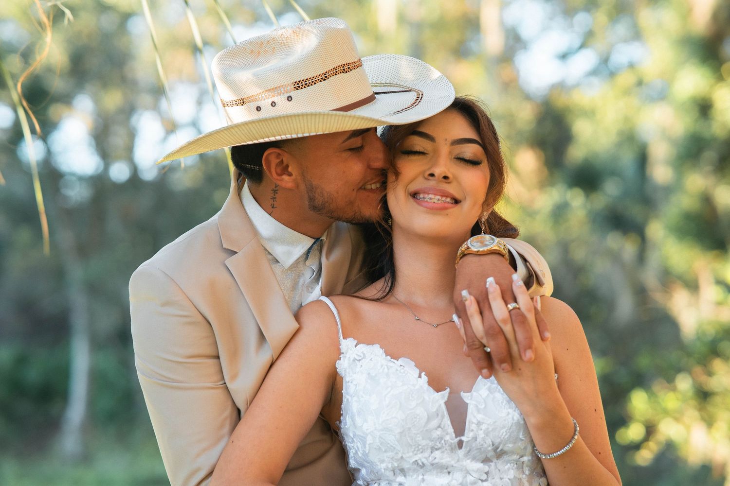 A romantic couple in western-style wedding attire share an intimate moment outdoors in warm sunlight.