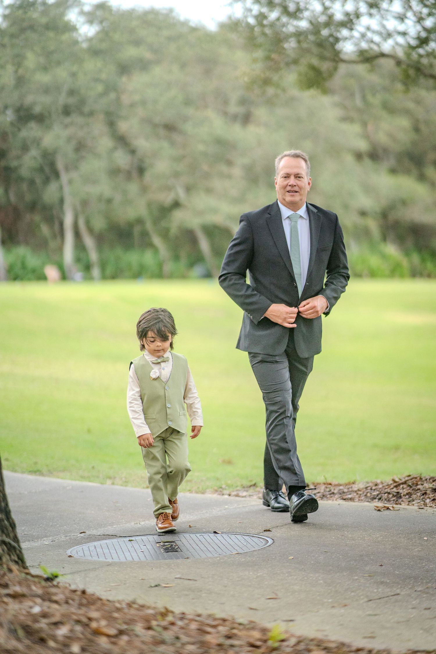 Two people in formal attire walk together on a path through a green park landscape on a sunny day.