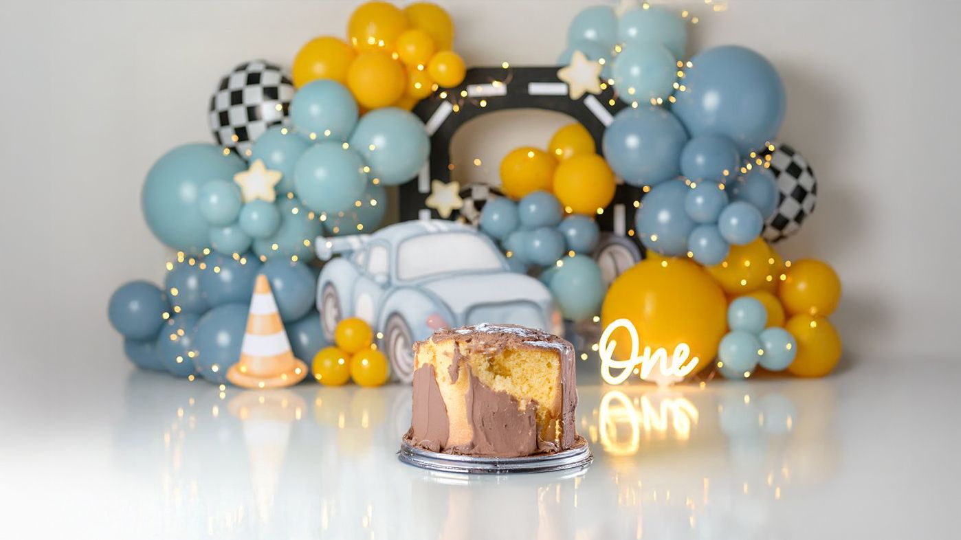 A birthday cake display with blue and yellow balloon decorations and a neon sign against a white backdrop.