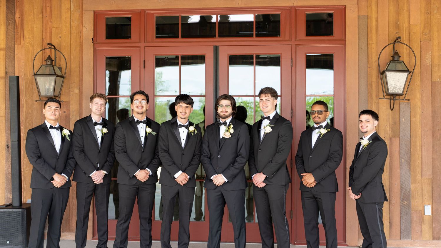 Groomsmen in black tuxedos with bow ties and boutonnieres pose outside rustic wooden venue doors.