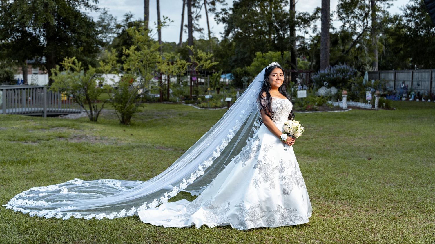 Elegant bride in a white ball gown with long lace veil posing gracefully outdoors on a sunny wedding day.