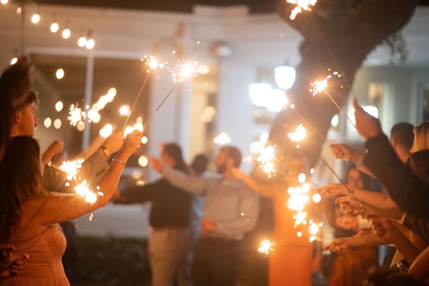People holding sparklers create a magical nighttime celebration atmosphere with glowing light trails in a warm indoor space.