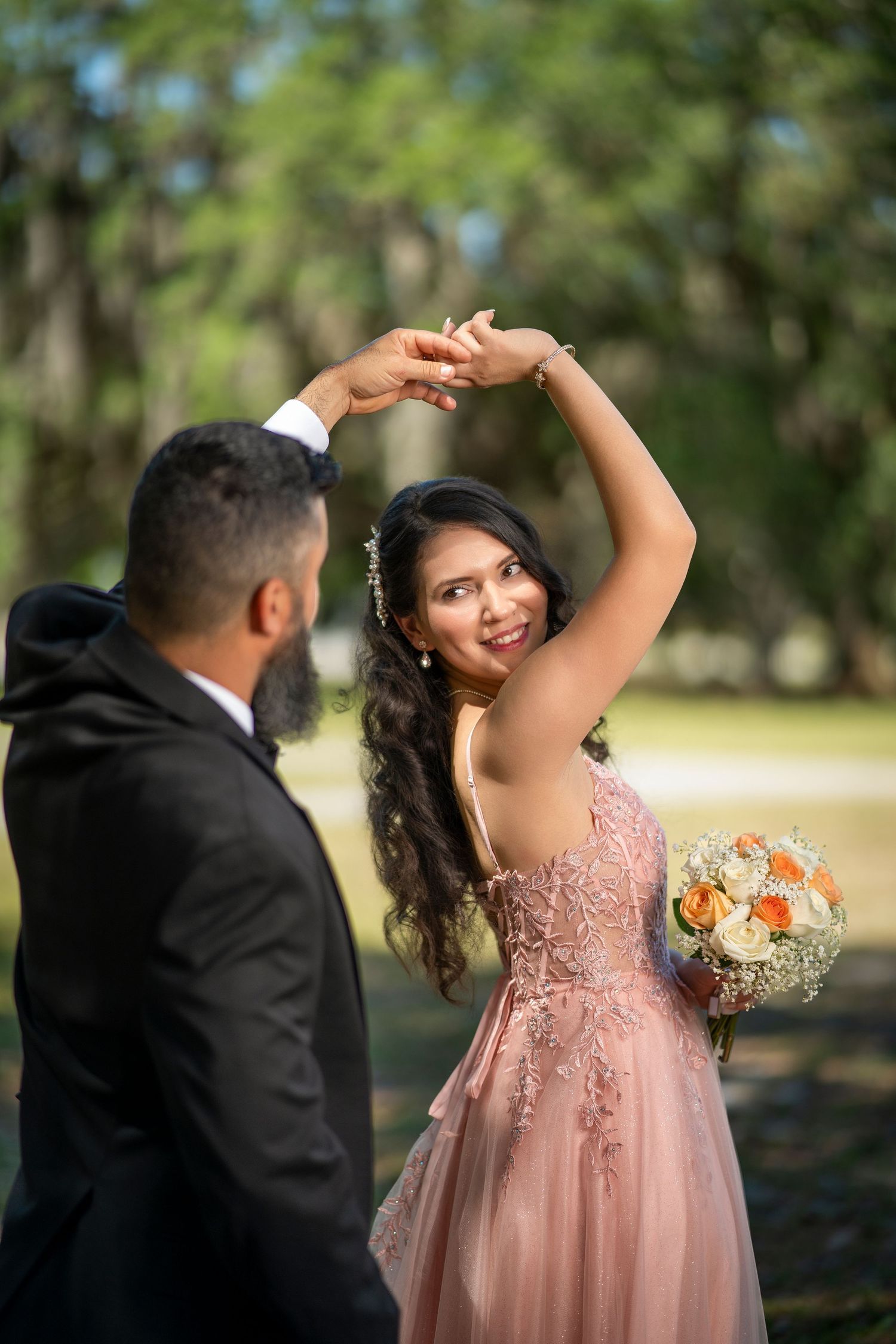 A bride in a pink lace gown holds a bouquet while dancing with a groom in a black suit outdoors.