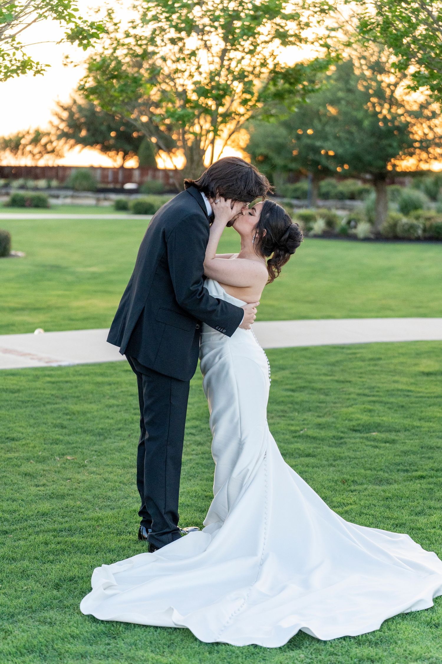 Bride and groom share romantic sunset kiss on lush green lawn in wedding attire.